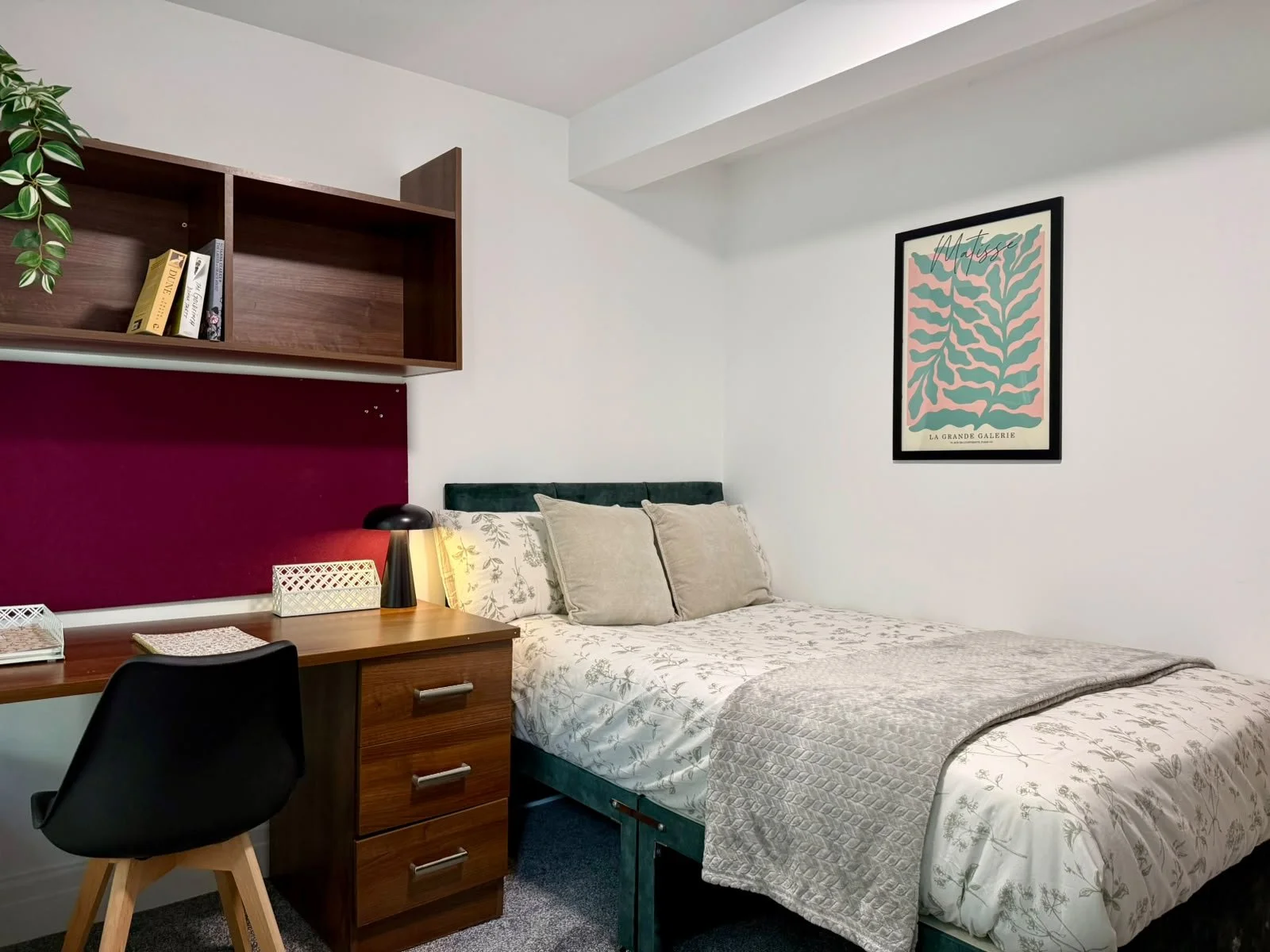 A cozy bedroom with a bed, cushions, a desk with a chair, a desk lamp, and a wall shelf with books. There is a framed art print on the wall above the bed.