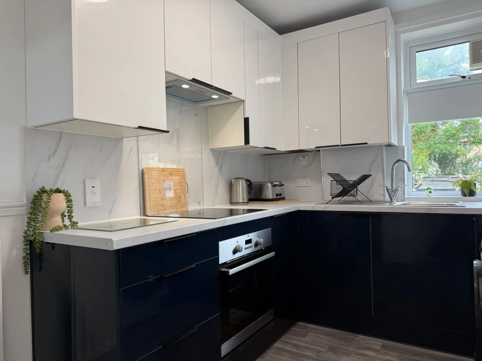 Modern kitchen with white and navy cabinets, marble backsplash, window with greenery outside, and various small appliances including toaster and kettle.