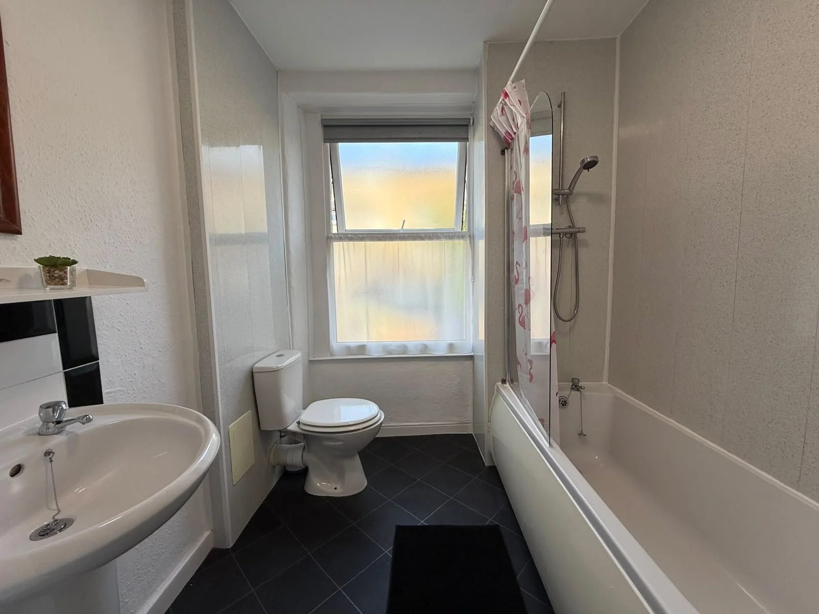 A small bathroom with a white sink, a toilet, a bathtub with a shower, a window with a frosted film, and a shower curtain with pink flamingos.