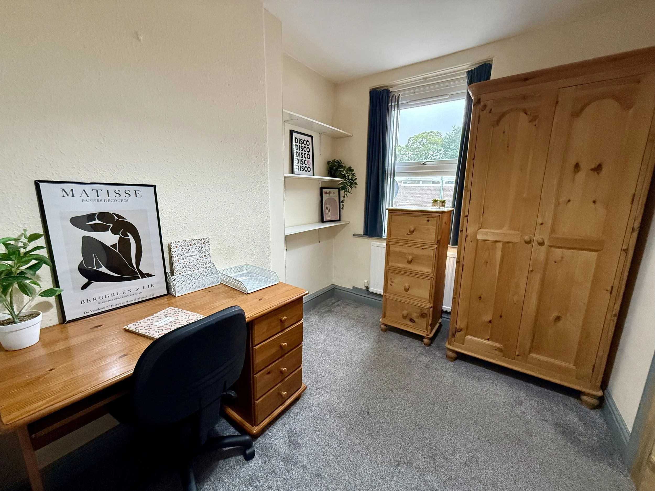 A small home office with a wooden desk, a black office chair, a potted plant, framed artwork on the desk, a bookshelf with decor, a window with blue curtains, and wooden furniture including a wardrobe and a chest of drawers.