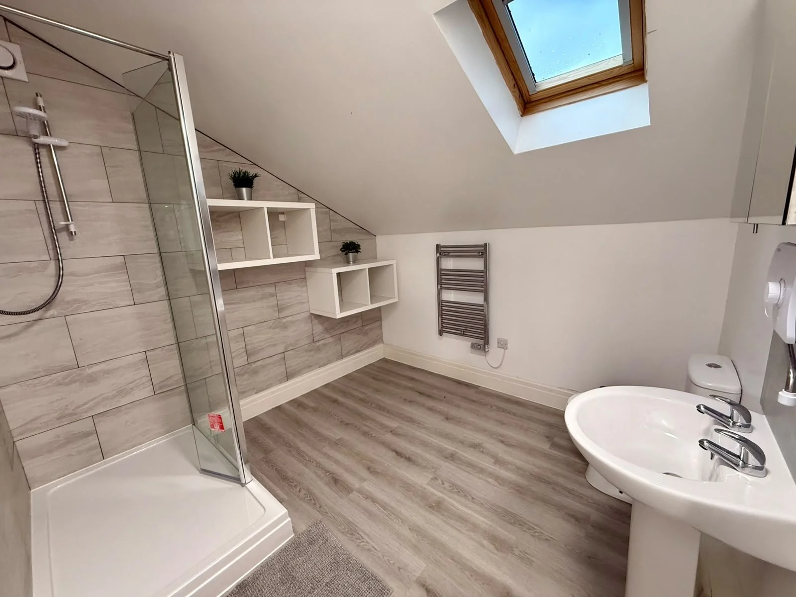 Modern bathroom with a shower enclosure, white shelves with potted plants, a window skylight, a towel radiator, a toilet, and a sink with dual faucets.