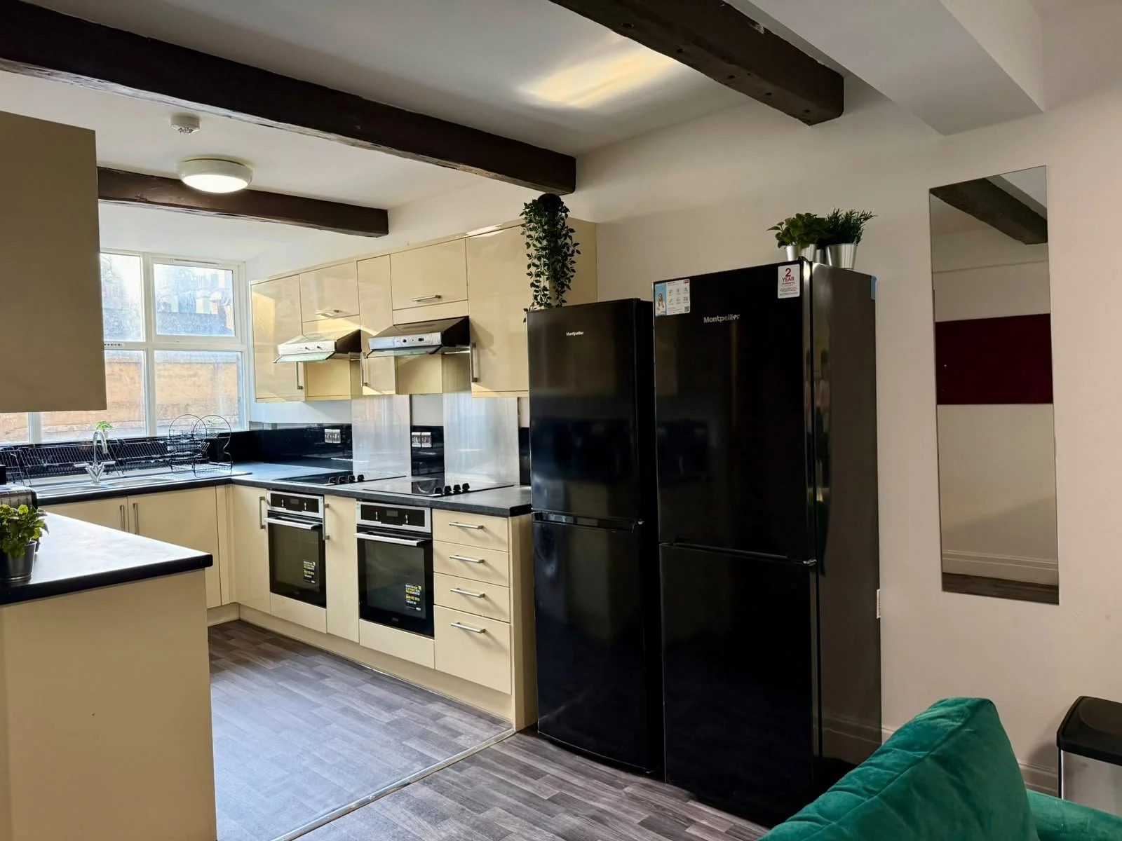 Modern kitchen with beige cabinets, black appliances, a window, potted plants, and wooden ceiling beams.