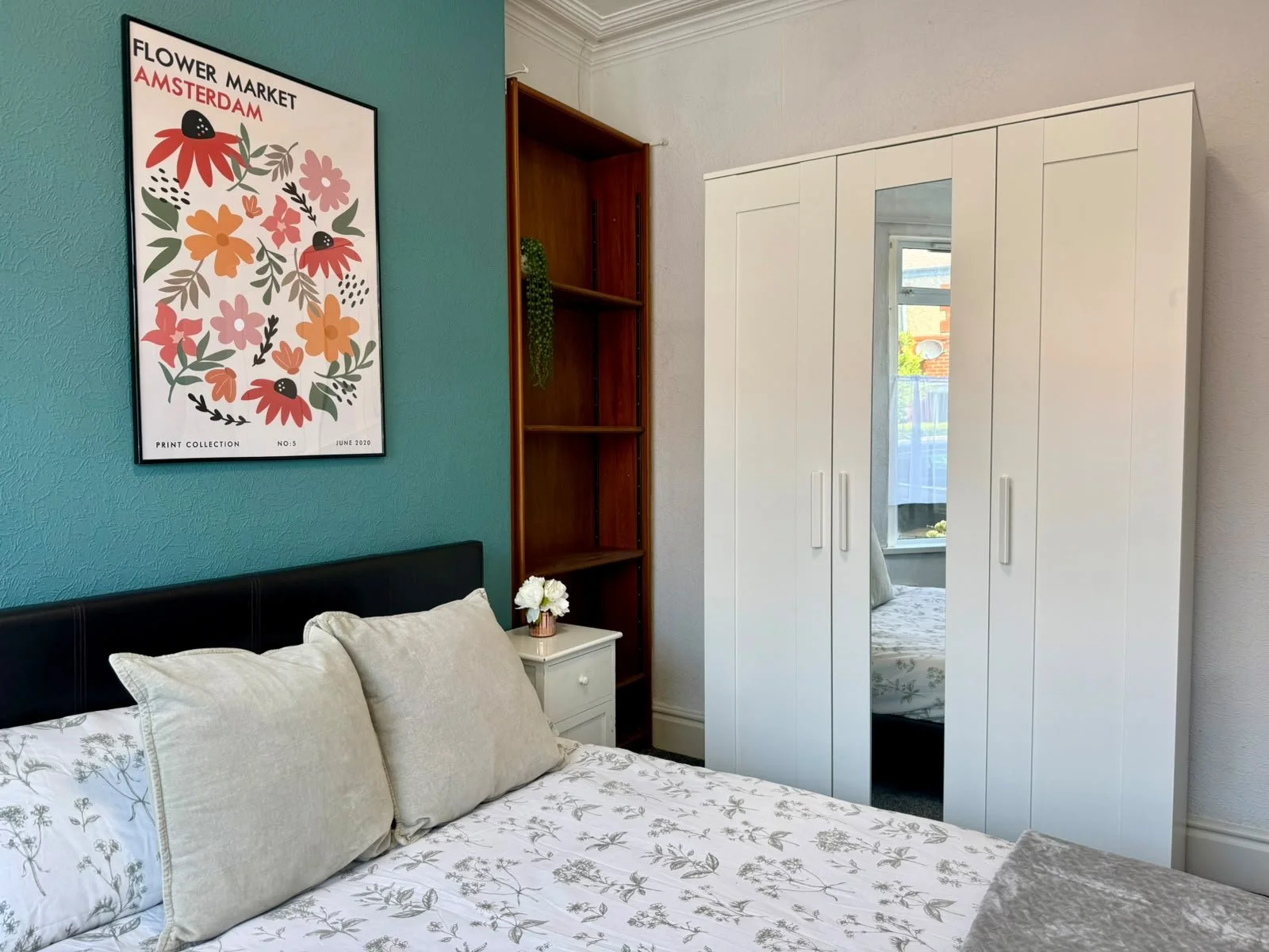 A bedroom corner with a bed, white pillows, and a side table with a small bouquet of white flowers. There's a large white wardrobe with a mirrored door, a wooden shelf unit, and a window in the background. A framed floral poster hangs on a teal wall.