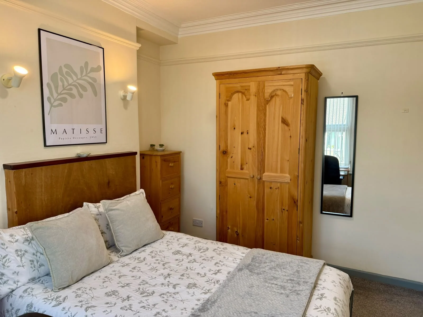 A bedroom with a bed, wooden headboard, pillows, a tall wooden wardrobe, small bedside drawers, a mirror on the wall, and wall-mounted lights, with a framed Matisse artwork hanging above the bed.