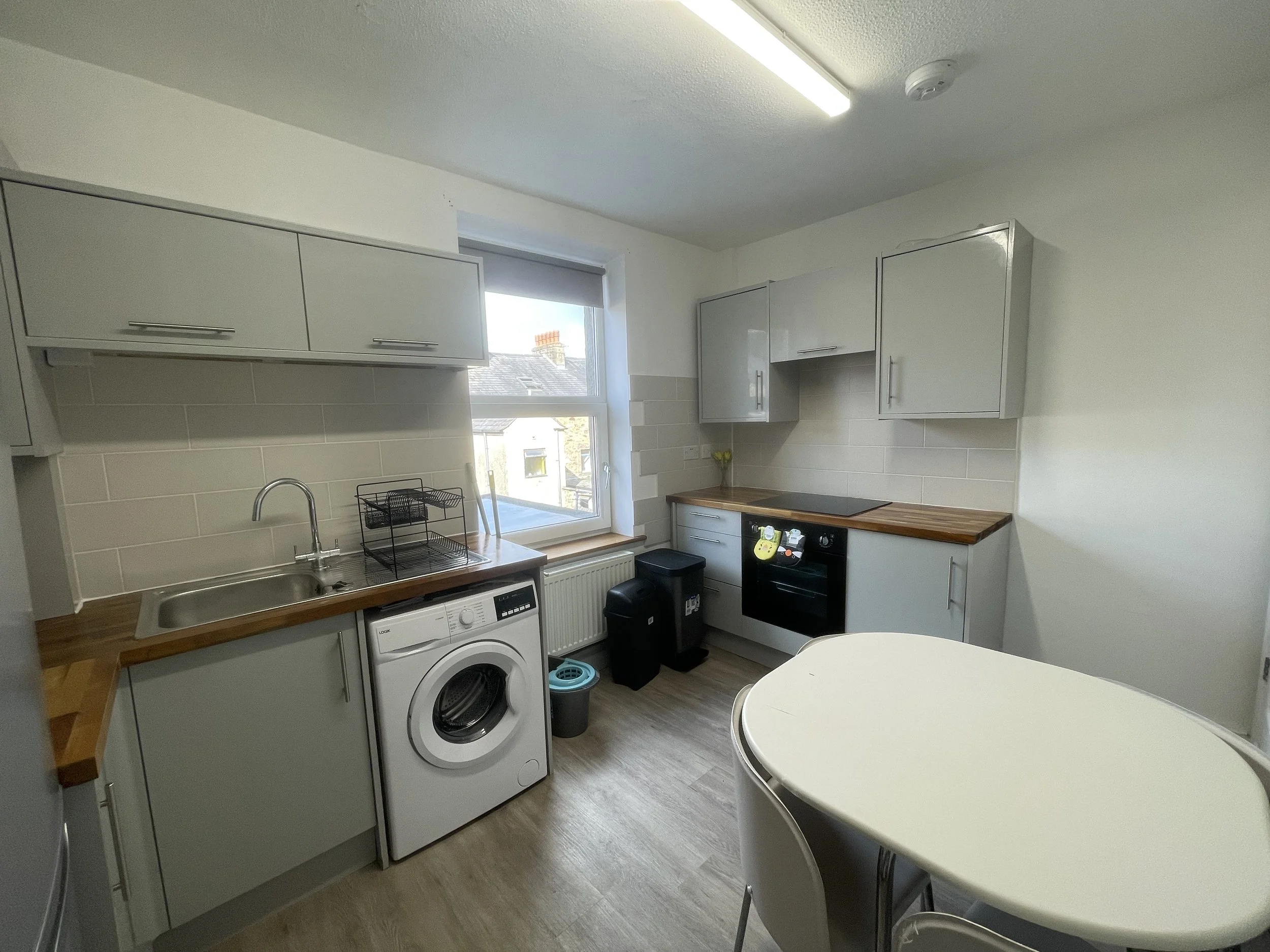 A small modern kitchen with light gray cabinets, a washing machine, a black oven, and a wooden countertop. There is a window with a view outside and a white table with chairs in the foreground.