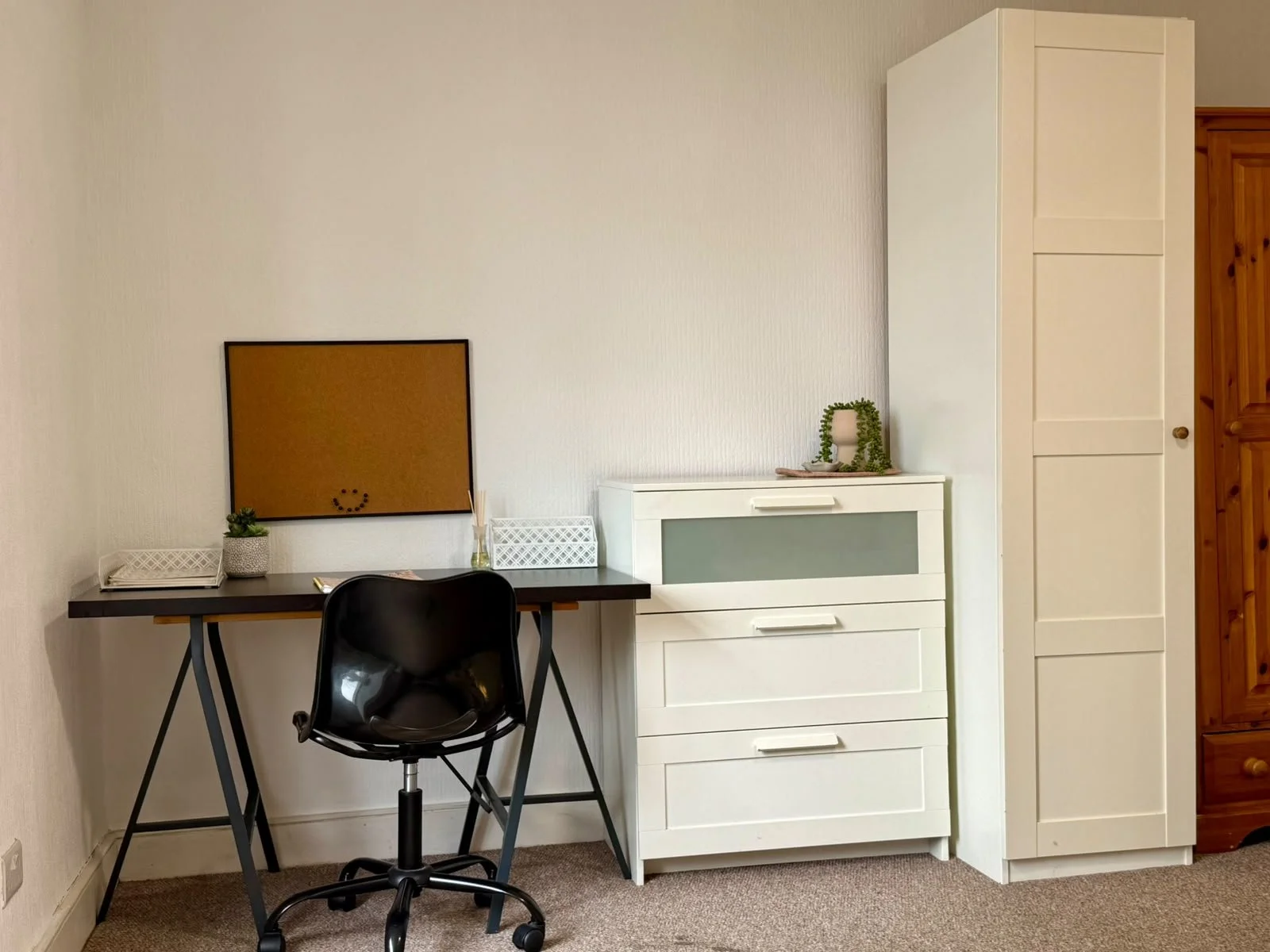 A small black desk with a black office chair, a small potted plant, and a tray with items on it. A corkboard on the wall, a white cabinet with drawers and a decorative plant on top, and a tall white wardrobe are in the background.