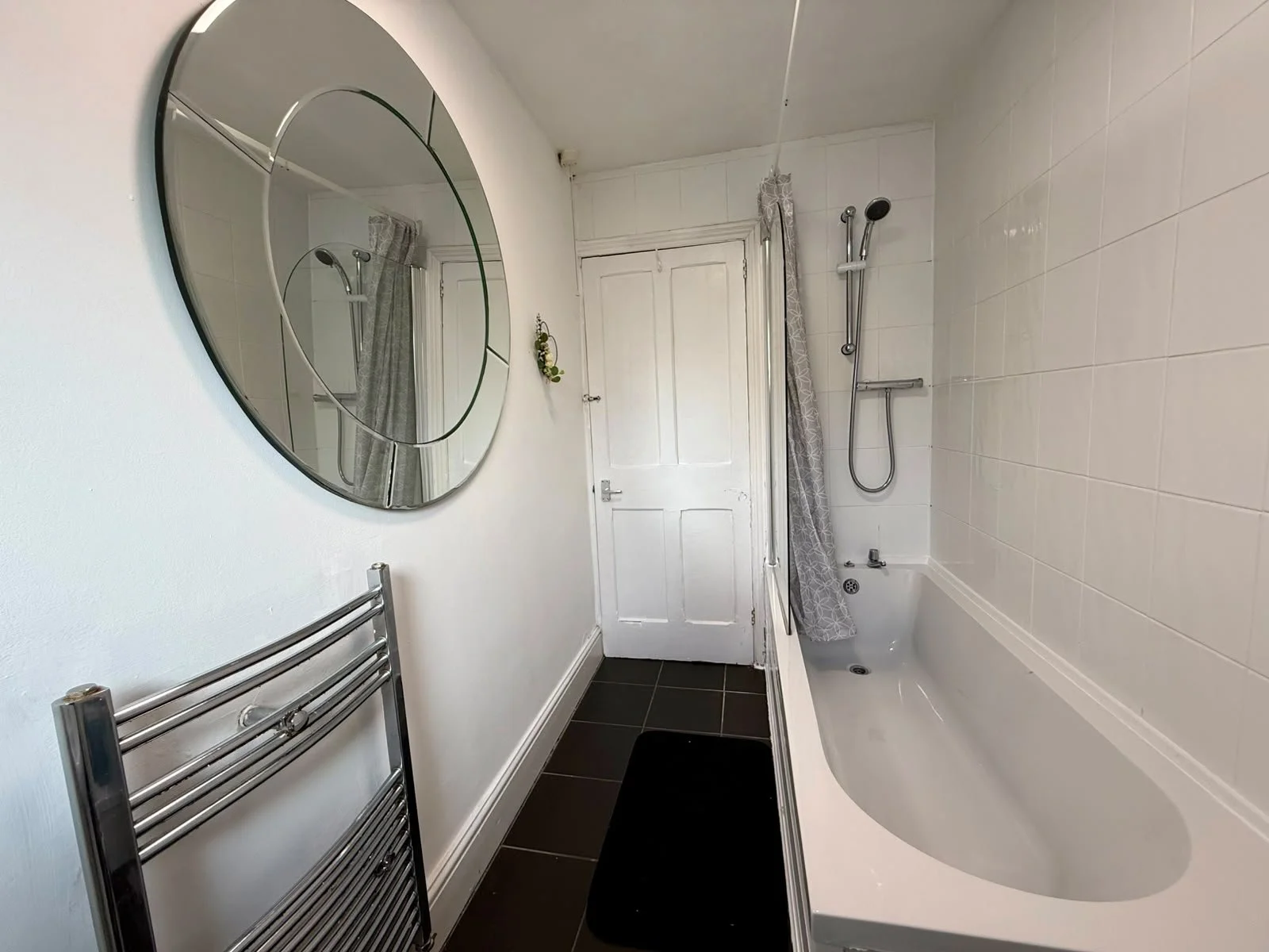 A bathroom featuring a wall-mounted mirror, a bathtub with a showerhead, a gray patterned shower curtain, a metal towel rack, and a white door.
