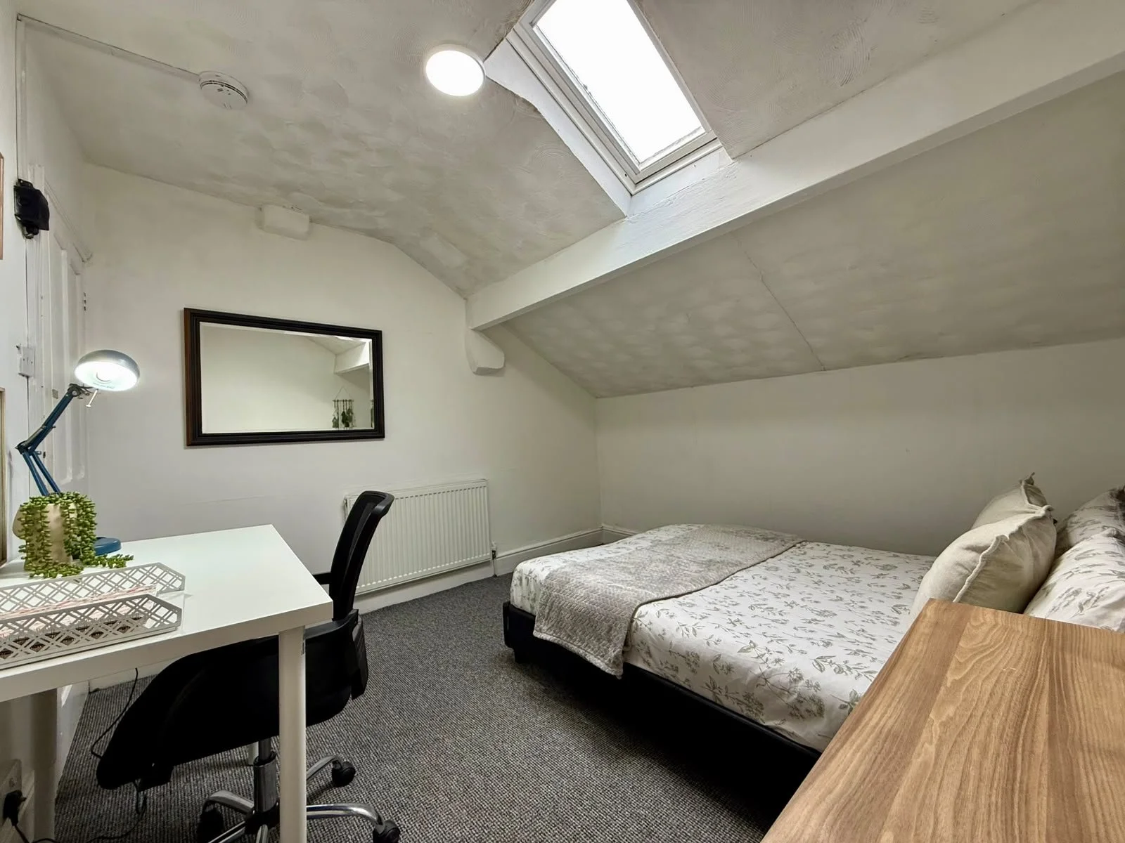 A cozy bedroom with a slanted ceiling and skylight. It features a bed with floral bedding, a white desk with a black office chair, a desk lamp, a mirror on the wall, and a small radiator. The room has white walls and a gray carpet.