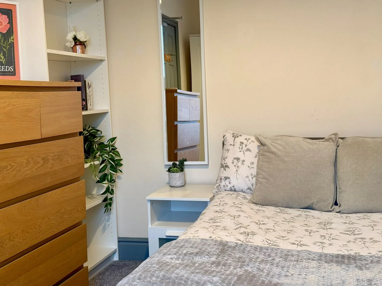 A cozy bedroom corner with a bed featuring patterned sheets and pillows, a white side table with a potted plant, a large mirror on the wall, a white bookshelf with decorative items and books, and a wooden dresser on the left side.