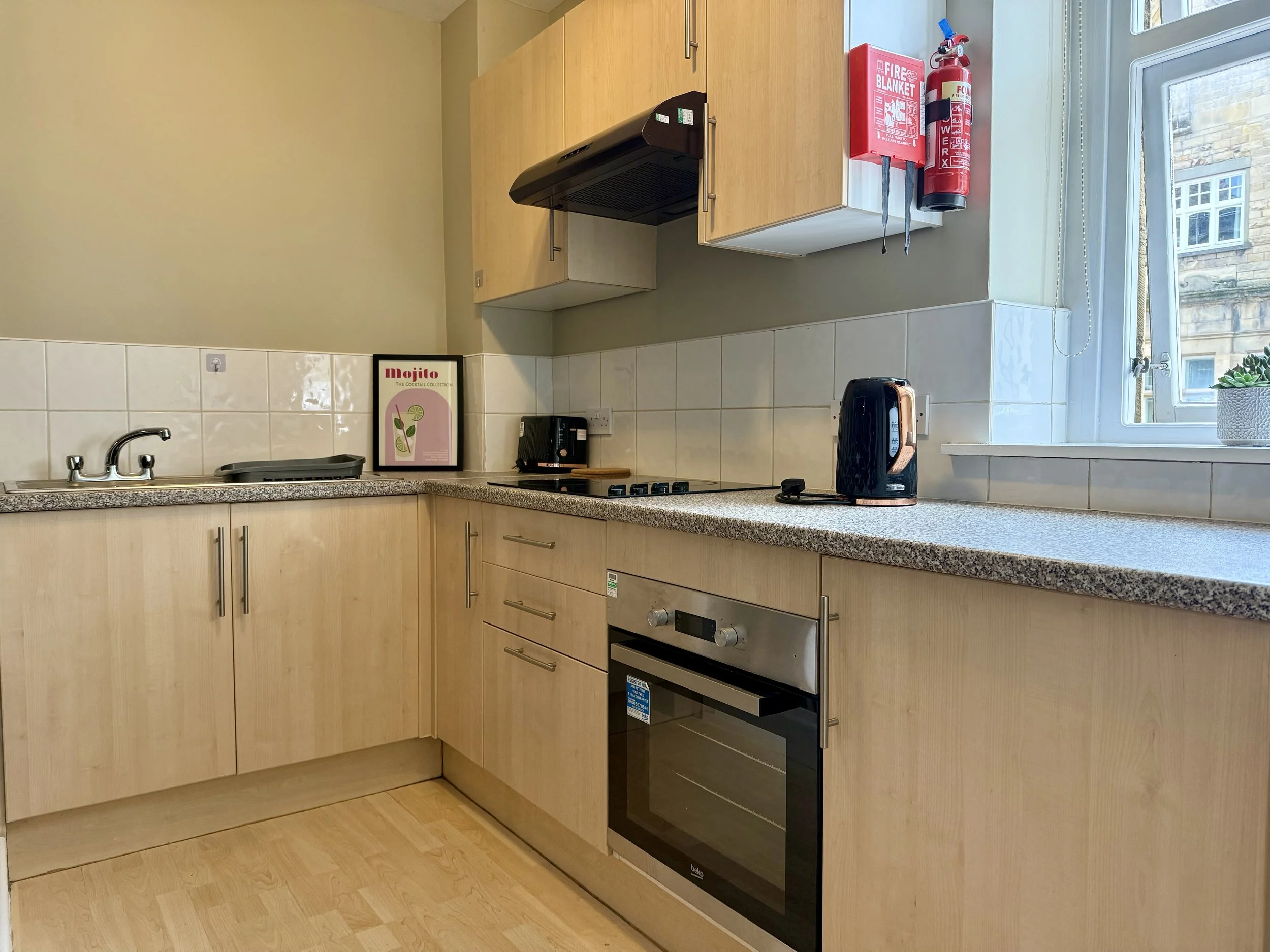 Kitchen with beige wooden cabinets and a speckled gray countertop. Items include a black kettle, a framed Mojito poster, a toaster, a gas stove, and a window with a small plant on the sill. Fire safety equipment such as a red fire blanket and fire ex