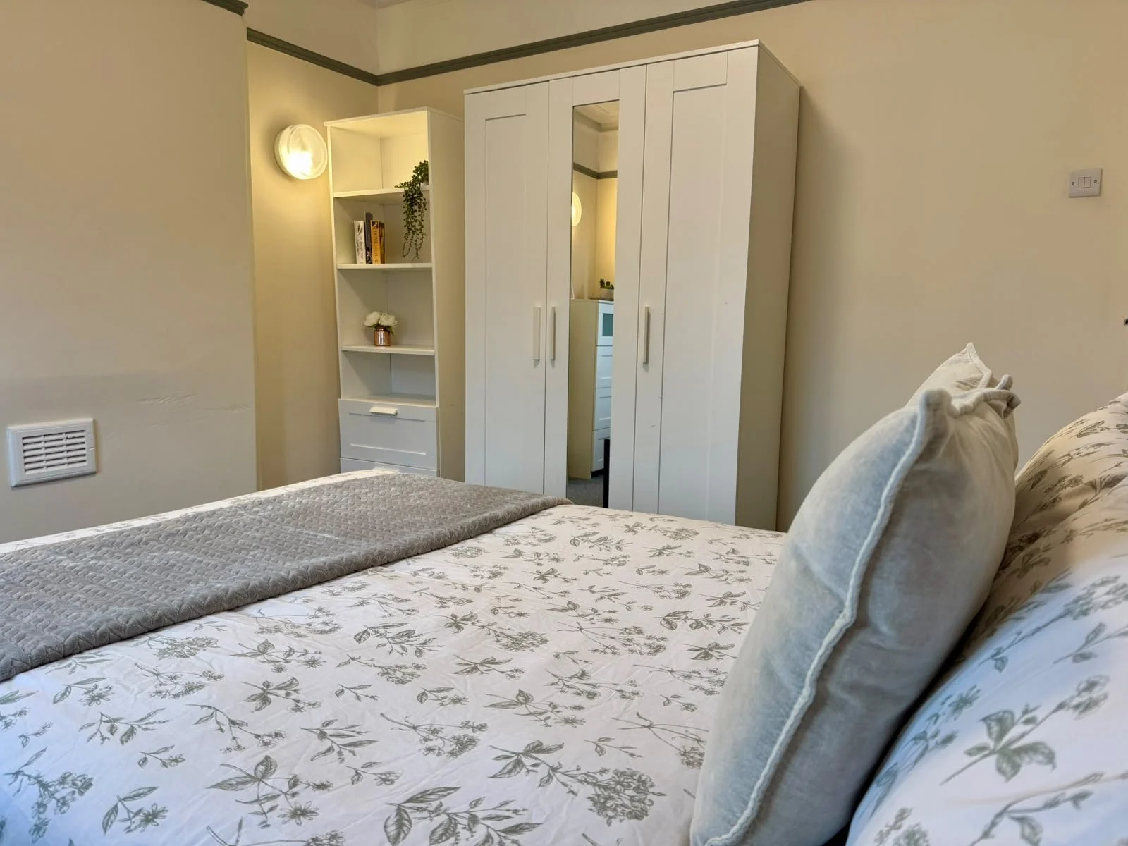 A neatly made bed with pillows and a floral bedspread in a bedroom with light-colored walls and built-in wardrobes.