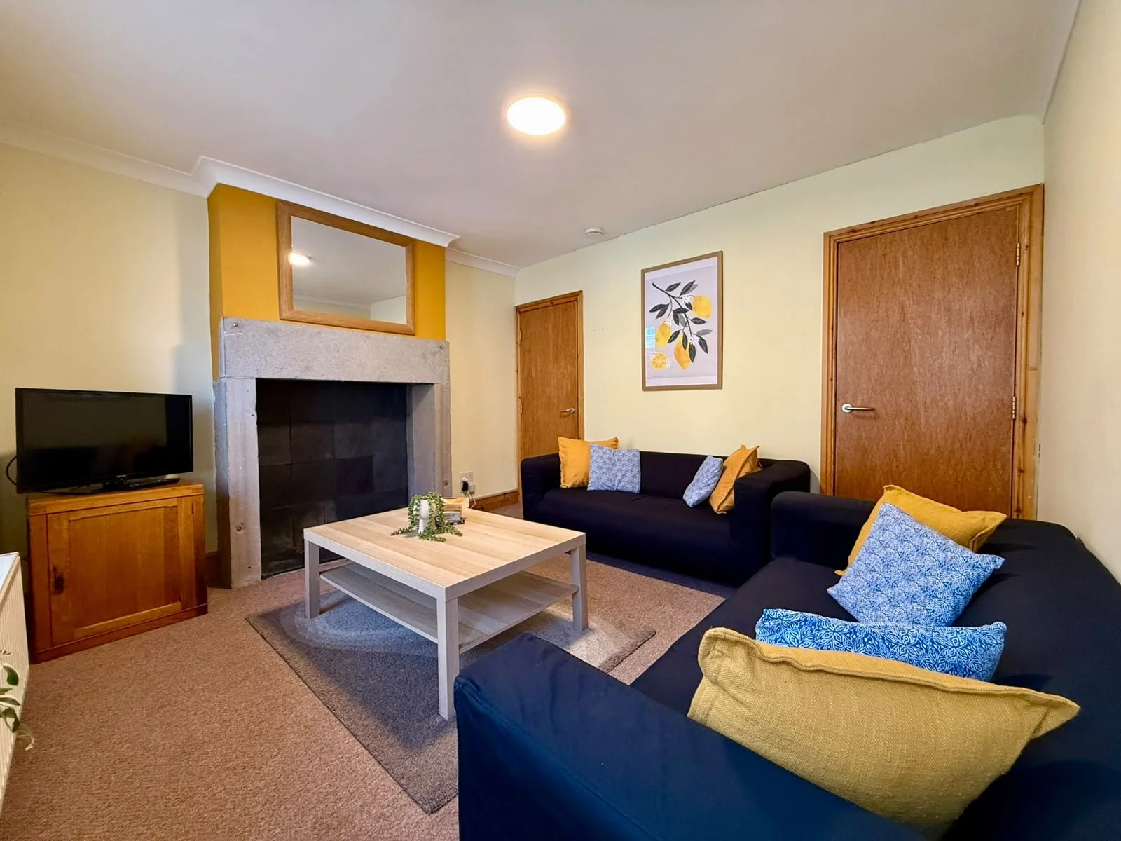 Living room with two black sofas with yellow and blue cushions, a light wooden coffee table, a stone fireplace with a mirror above, a small wooden TV stand with a TV, and a framed art print of lemons on a pale yellow wall.