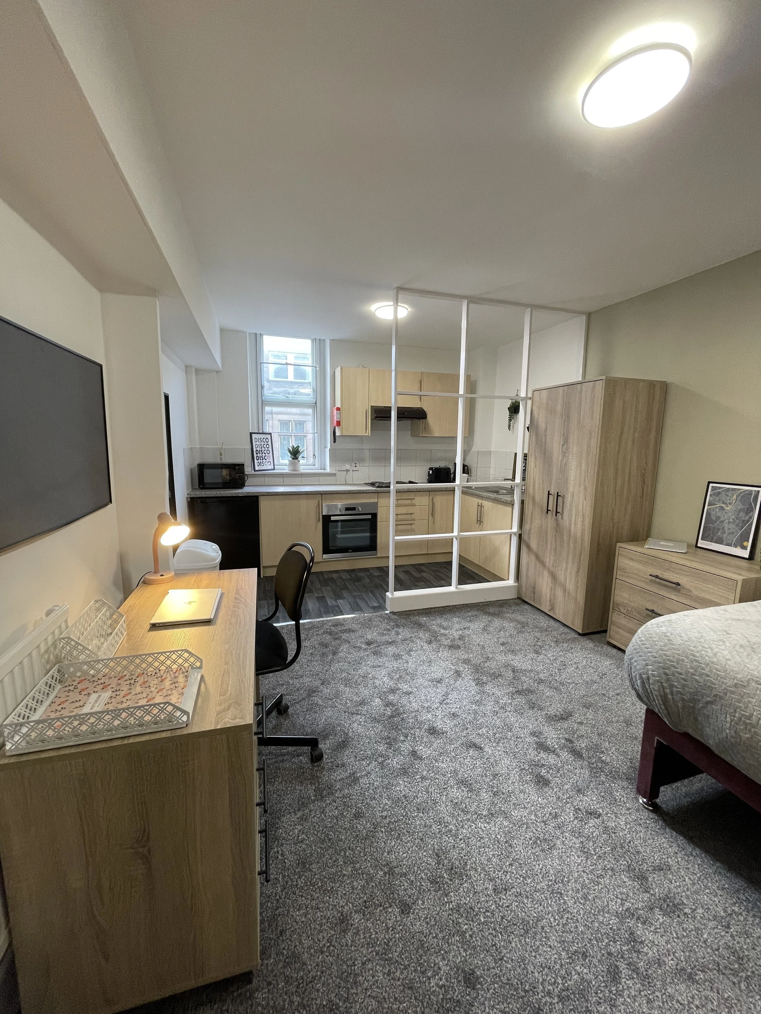 A small, modern apartment with a combined living, kitchen, and workspace area. The room includes a desk with a lamp, a chair, a bed, a wardrobe, and a cabinet. The kitchen features light wood cabinets, appliances, and a window with a small plant and