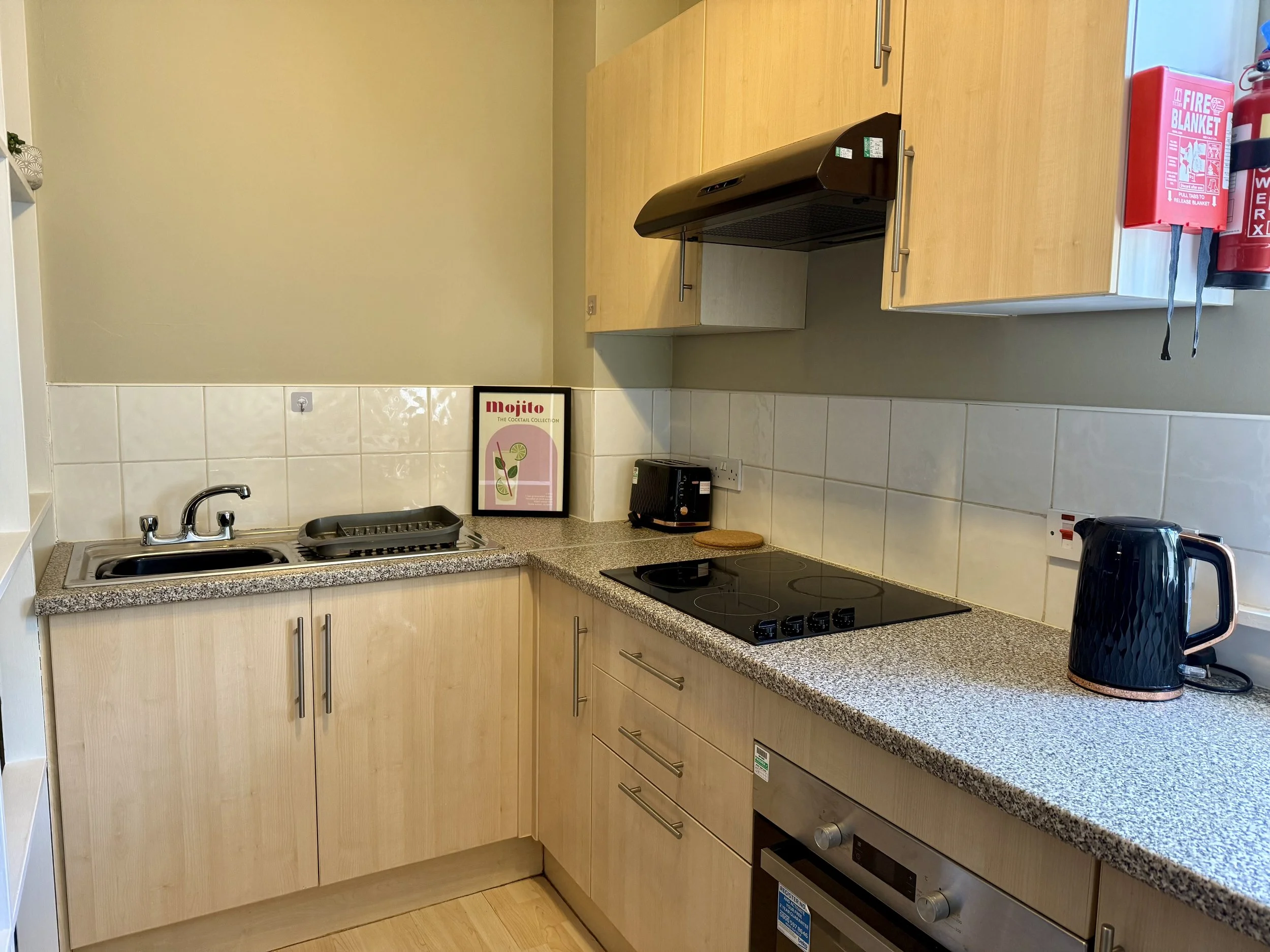 Kitchen with light wood cabinets, granite countertop, black stovetop, black kettle, and framed mojito poster near the sink.