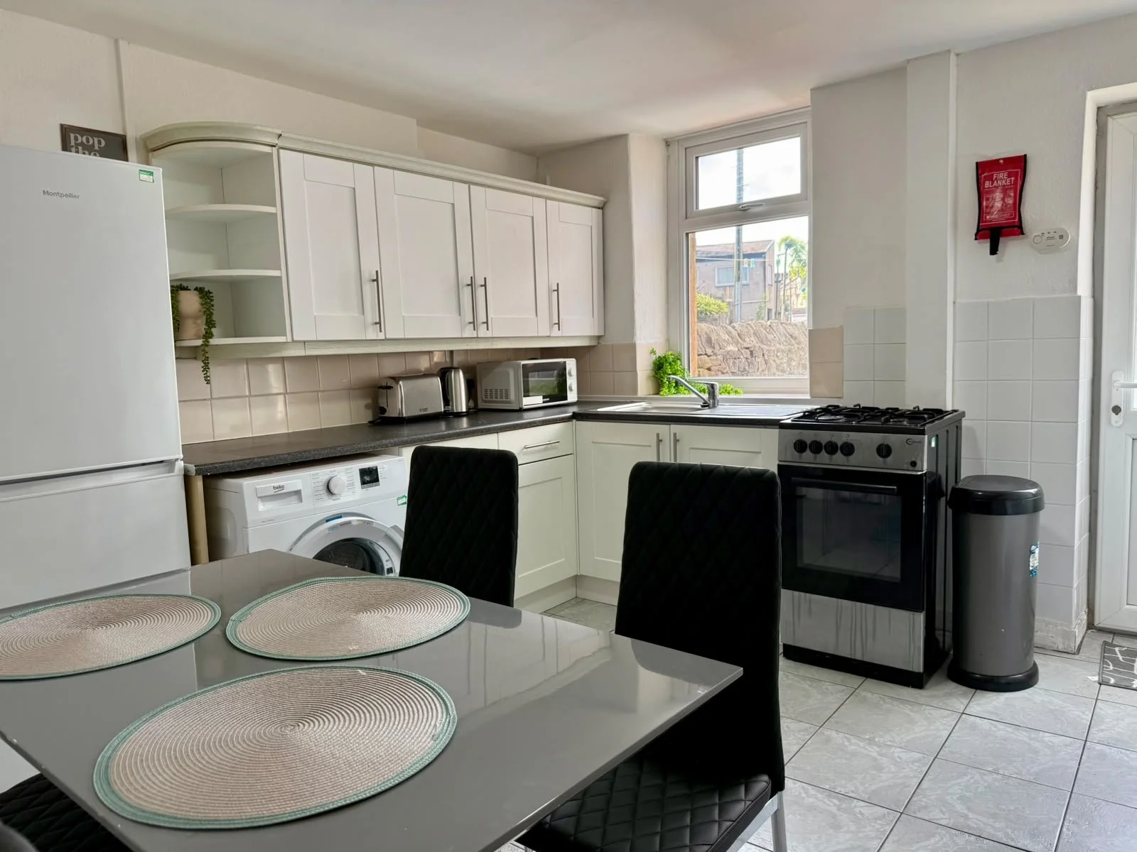 Kitchen with white cabinets, black countertop, and appliances including a refrigerator, washing machine, microwave, toaster, kettle, and stove. There are two black chairs, a white dining table with placemats, and a window with a view outside.