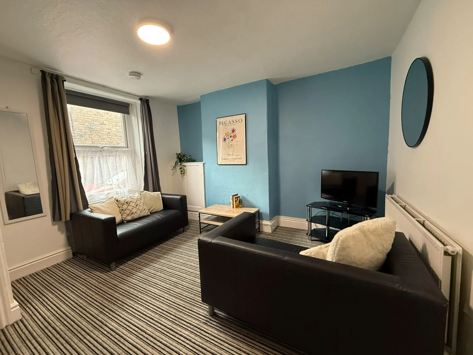 Living room with two black leather sofas, striped carpet, small TV on a black stand, blue accent wall with art poster, window with curtains, radiator, and mirror.