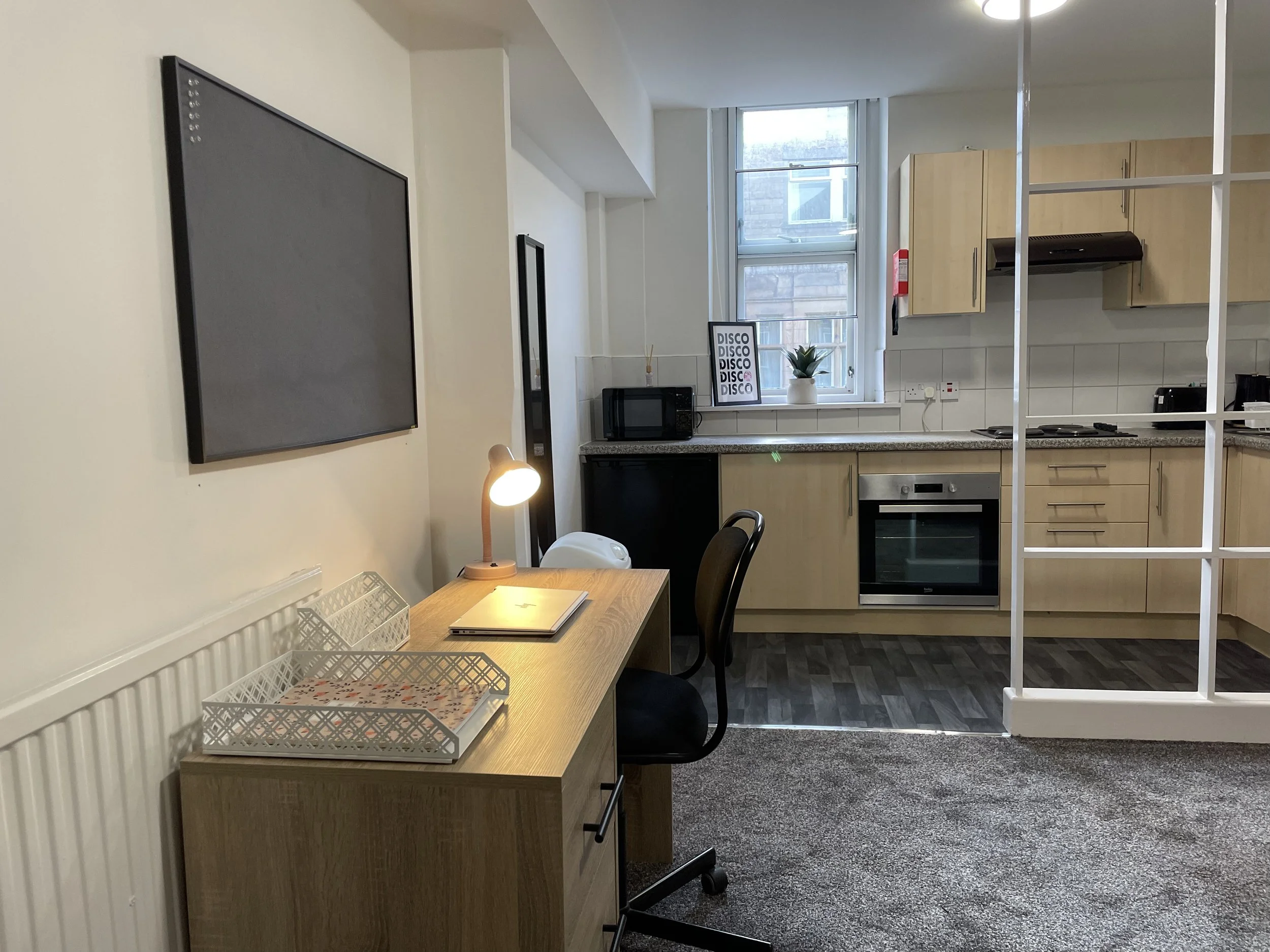 A compact apartment kitchen and workspace with a small desk, chair, and a lamp, along with a kitchen area featuring light wood cabinets, a microwave, stove, oven, and a window with natural light.