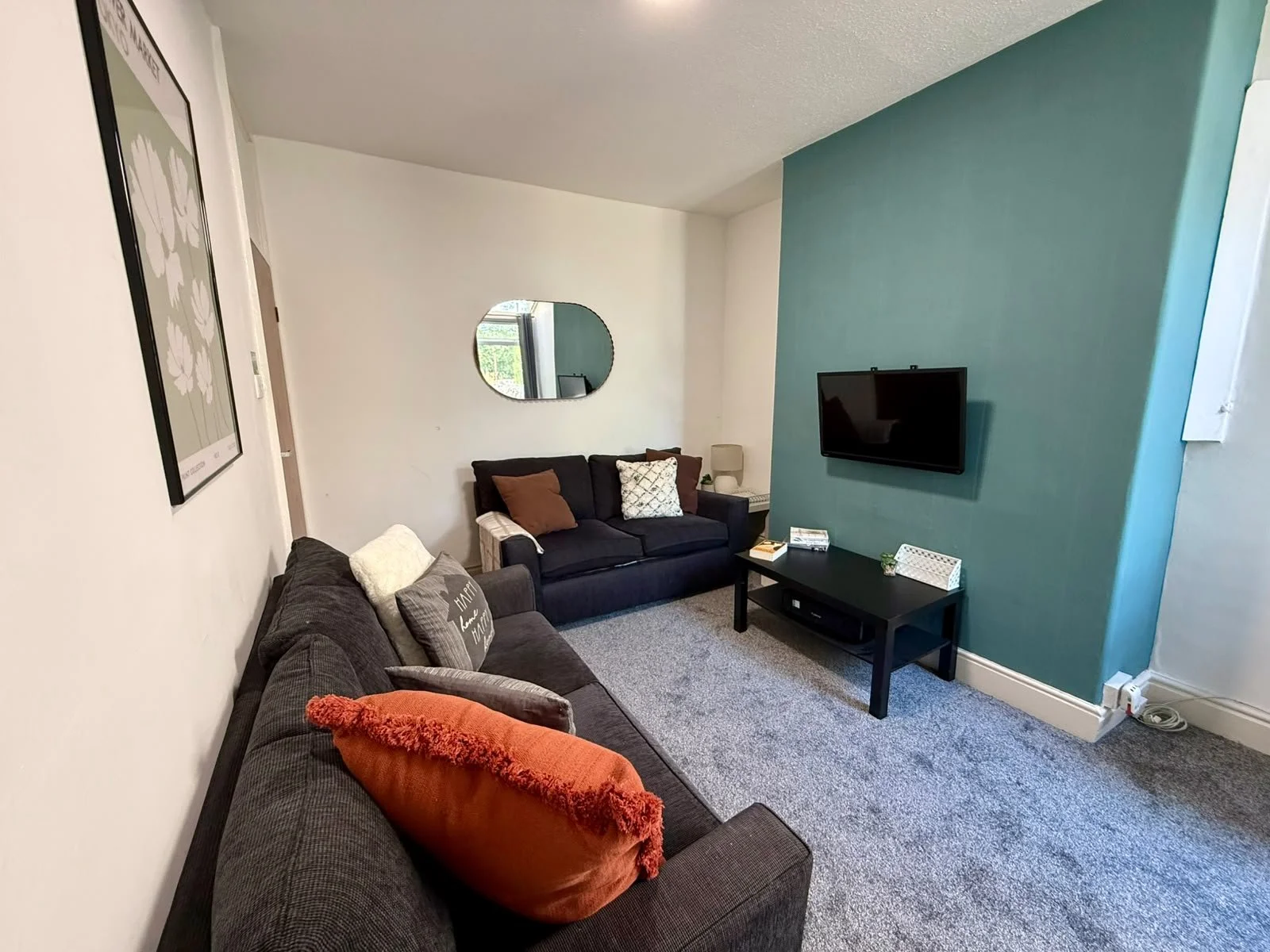Living room with gray carpet, dark gray and black sofas, orange and patterned pillows, a black coffee table, a wall-mounted TV, a teal accent wall, white walls, a small side table with a lamp, mirror, and framed art.