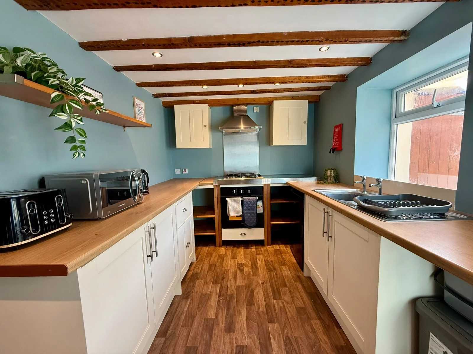 A modern kitchen with wooden countertops, white cabinets, a stainless steel range hood, and a blue accent wall. There is a microwave, toasters, and a sink with a drying rack. Wooden ceiling beams and a window bring natural light.
