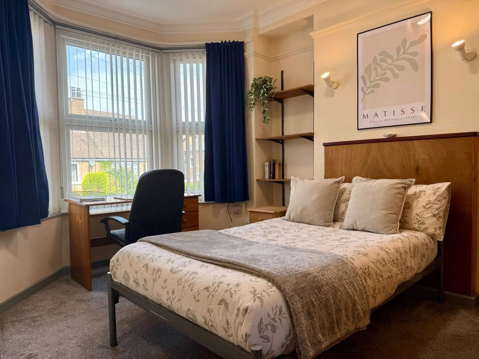 A bedroom with a large bed, beige floral bedding, and pillows. There are blue and white curtains, a window looking outside, a desk with a chair, and wall shelves with books. A framed art print of a leaf titled 'Maïsse' hangs above the bed.