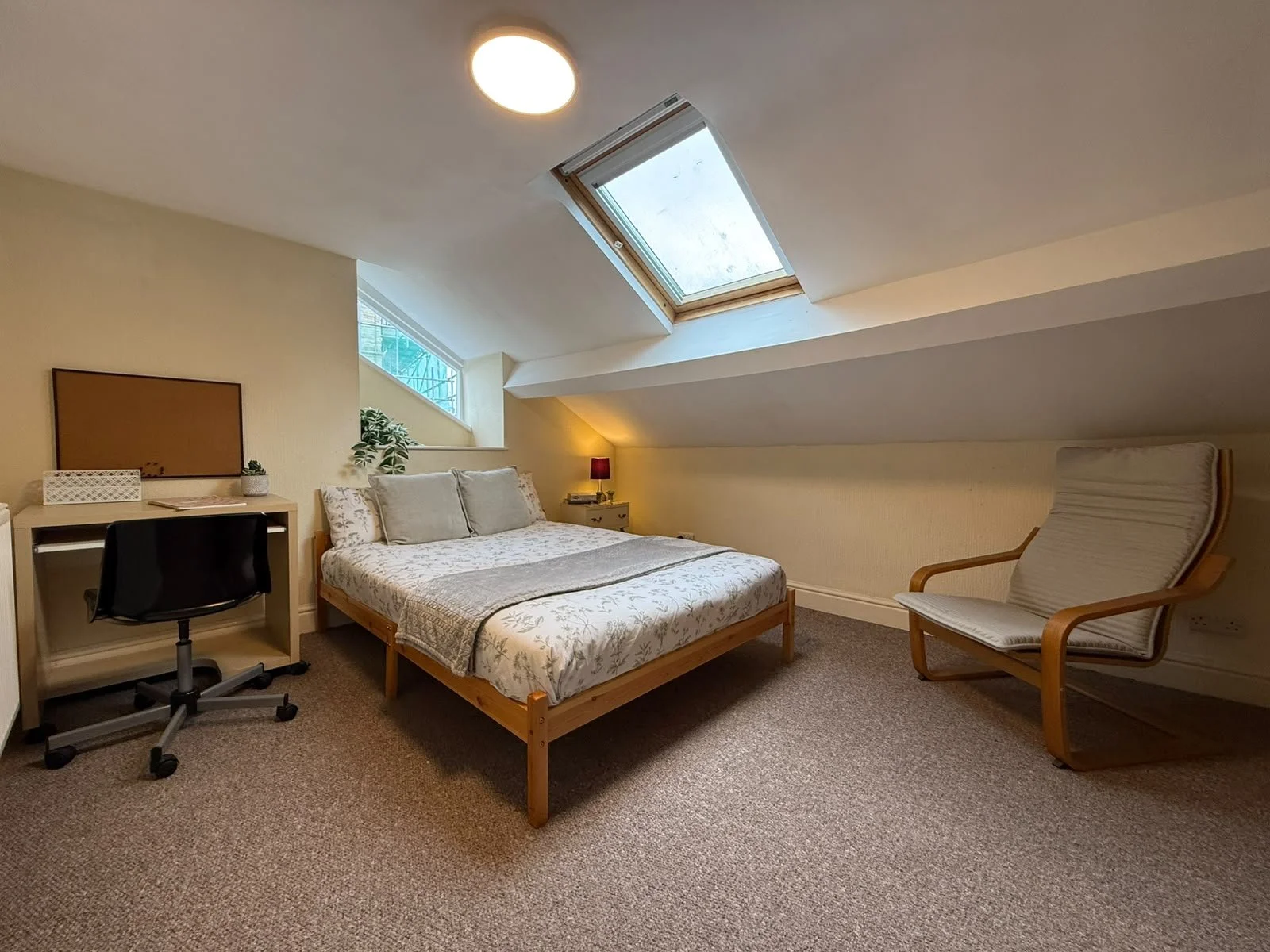 A bedroom with a bed, desk, chair, armchair, and skylight window in an attic space.