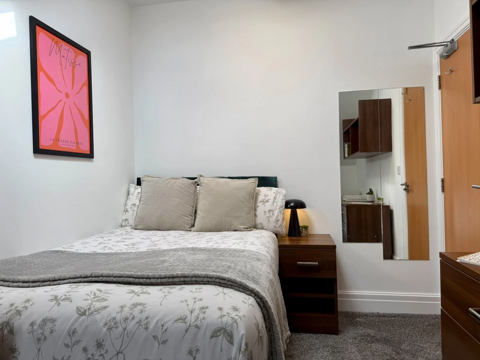 A bedroom with a bed that has a gray quilt and floral bedding, three beige pillows, a wooden nightstand with a black lamp and a small plant, a mirror on the wall, and a framed pink poster on the wall.