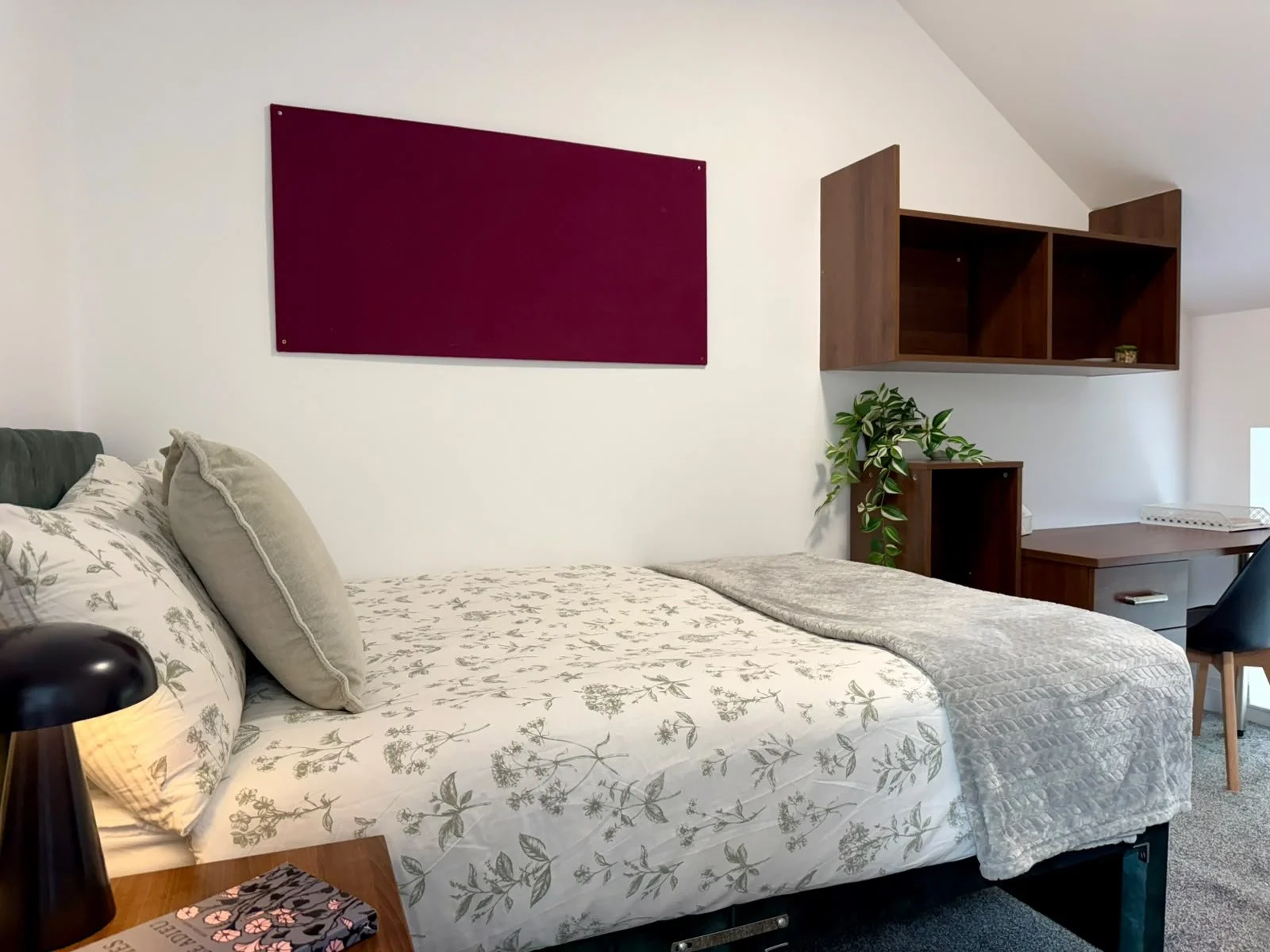 Bedroom with a bed having floral bedspread and beige pillows, a black bedside lamp, a wooden desk with a drawer, a navy blue chair, a potted plant, and wooden wall shelves, with a purple wall decoration on the wall.