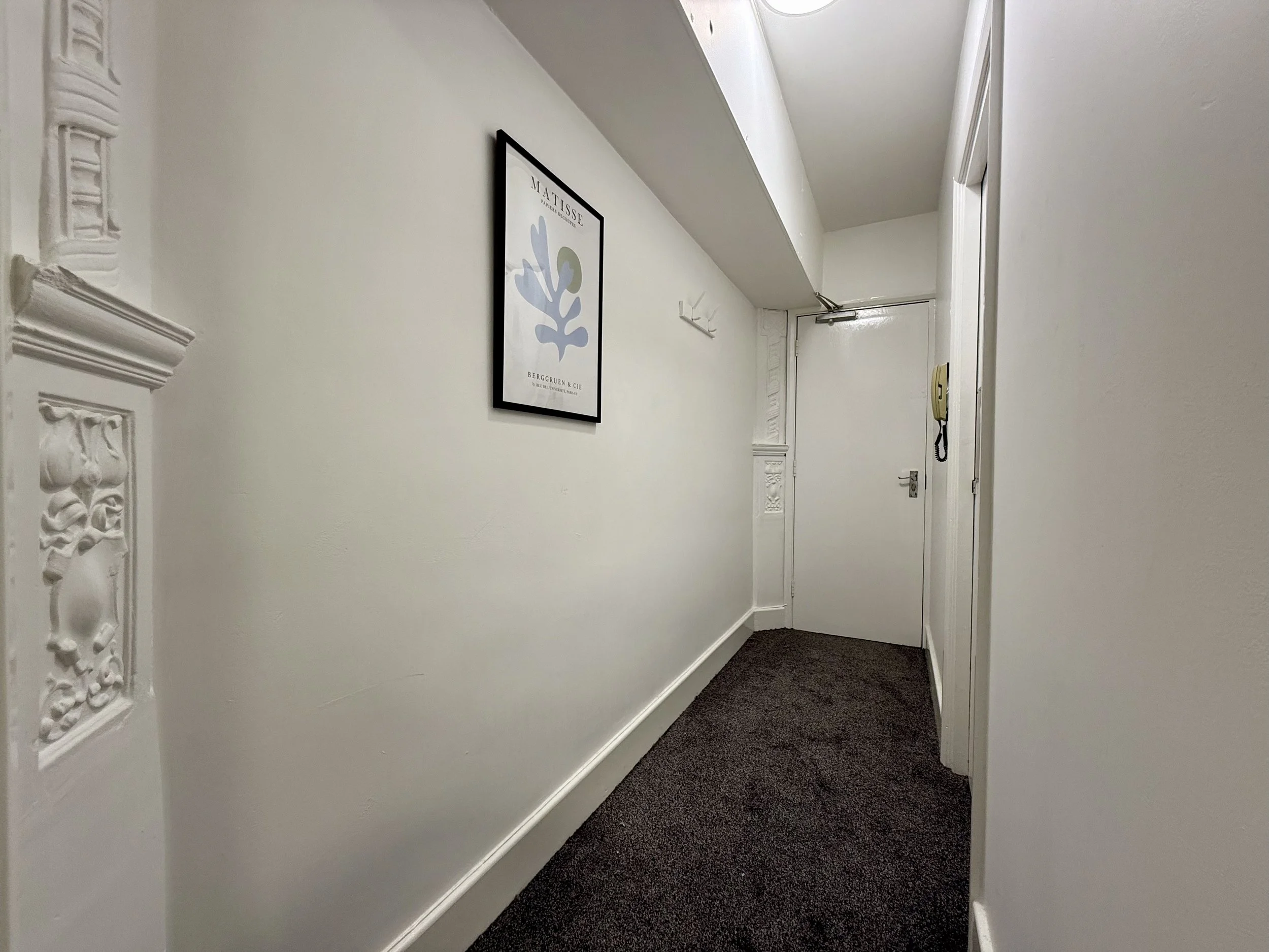 A narrow apartment hallway with white walls, a brown carpet, and decorative molding on the wall. A framed poster of Matisse is hanging on the wall. There is a white door at the end of the hallway and a beige intercom on the right wall.
