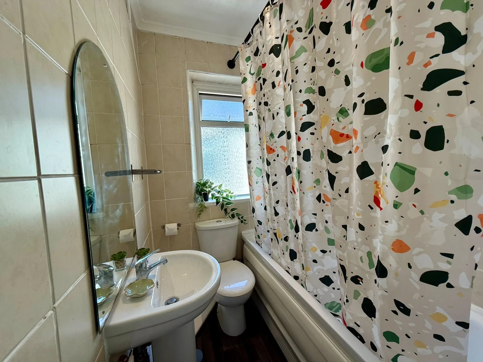 Small bathroom with a window, a white toilet, a sink with a mirror, a patterned shower curtain, and a potted plant on the windowsill.