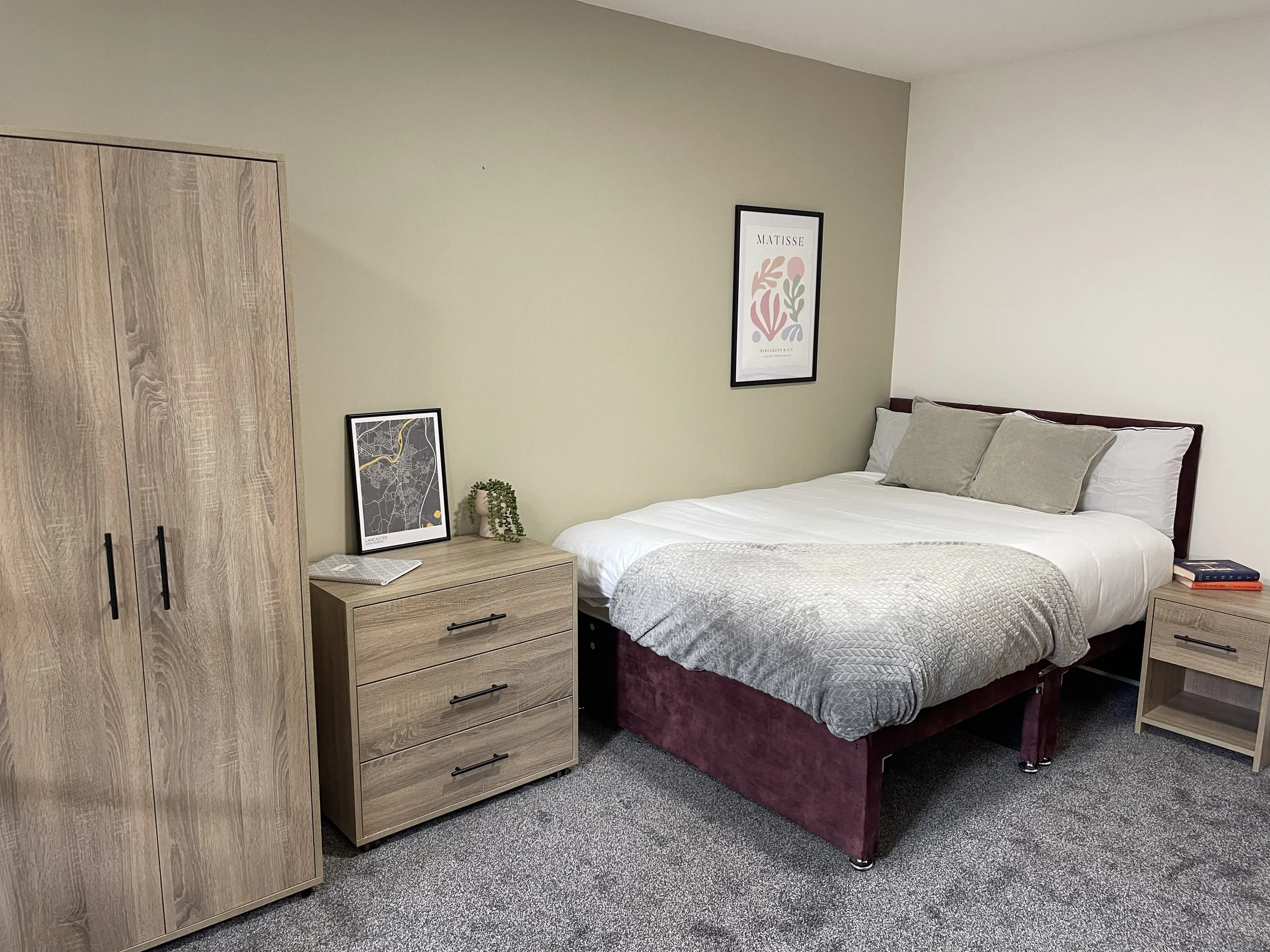 A cozy bedroom with a bed, nightstands, a wardrobe, and decor. The bed has white and beige bedding with three pillows. A framed poster hangs on the wall above the bed. There is a small plant and framed picture on the left nightstand, and books on the
