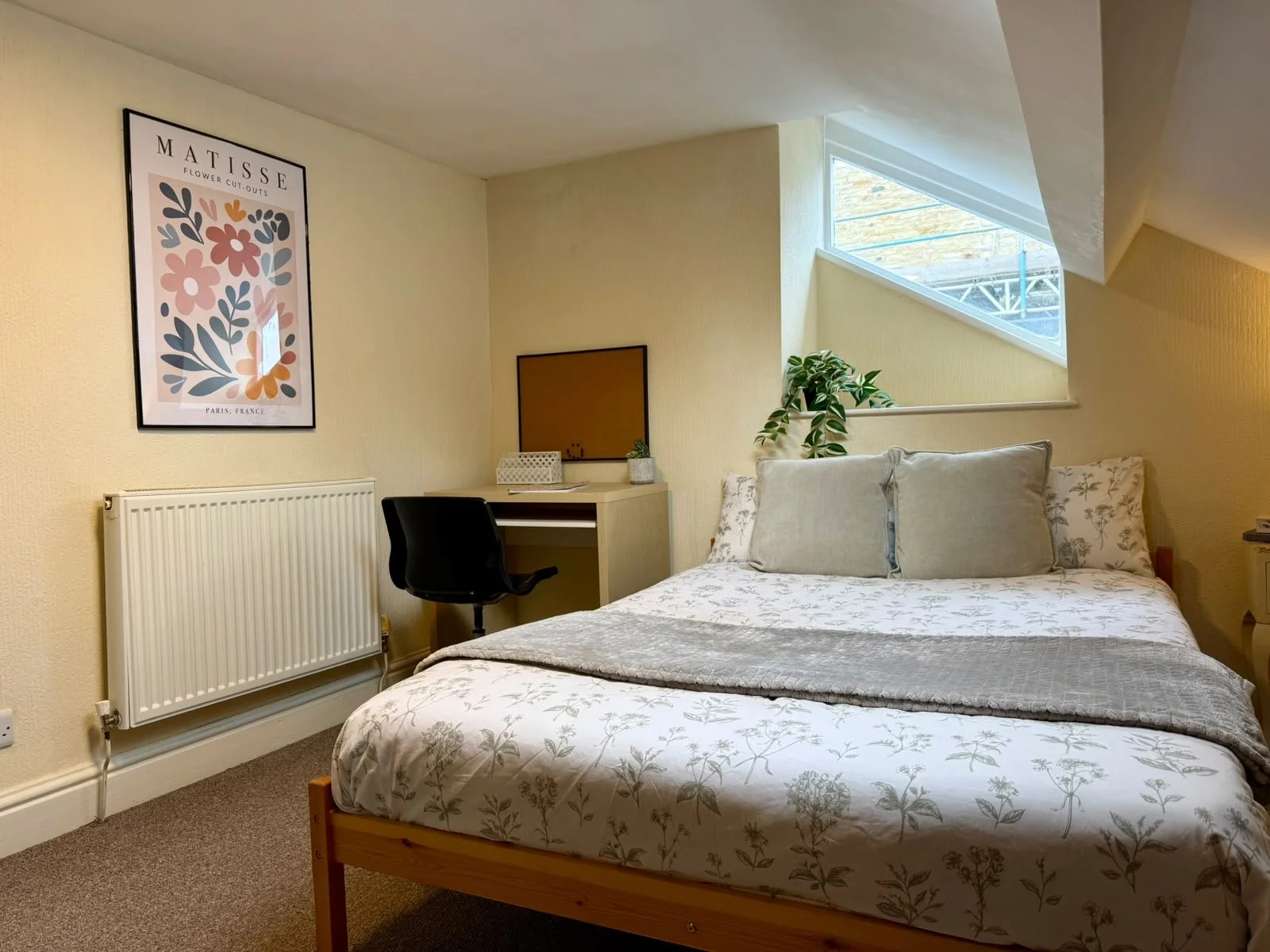 A cozy bedroom with a double bed, decorative pillows, a wooden headboard, a small desk with a chair, a potted plant, a radiator, and framed wall art, illuminated by natural light from a triangular window.