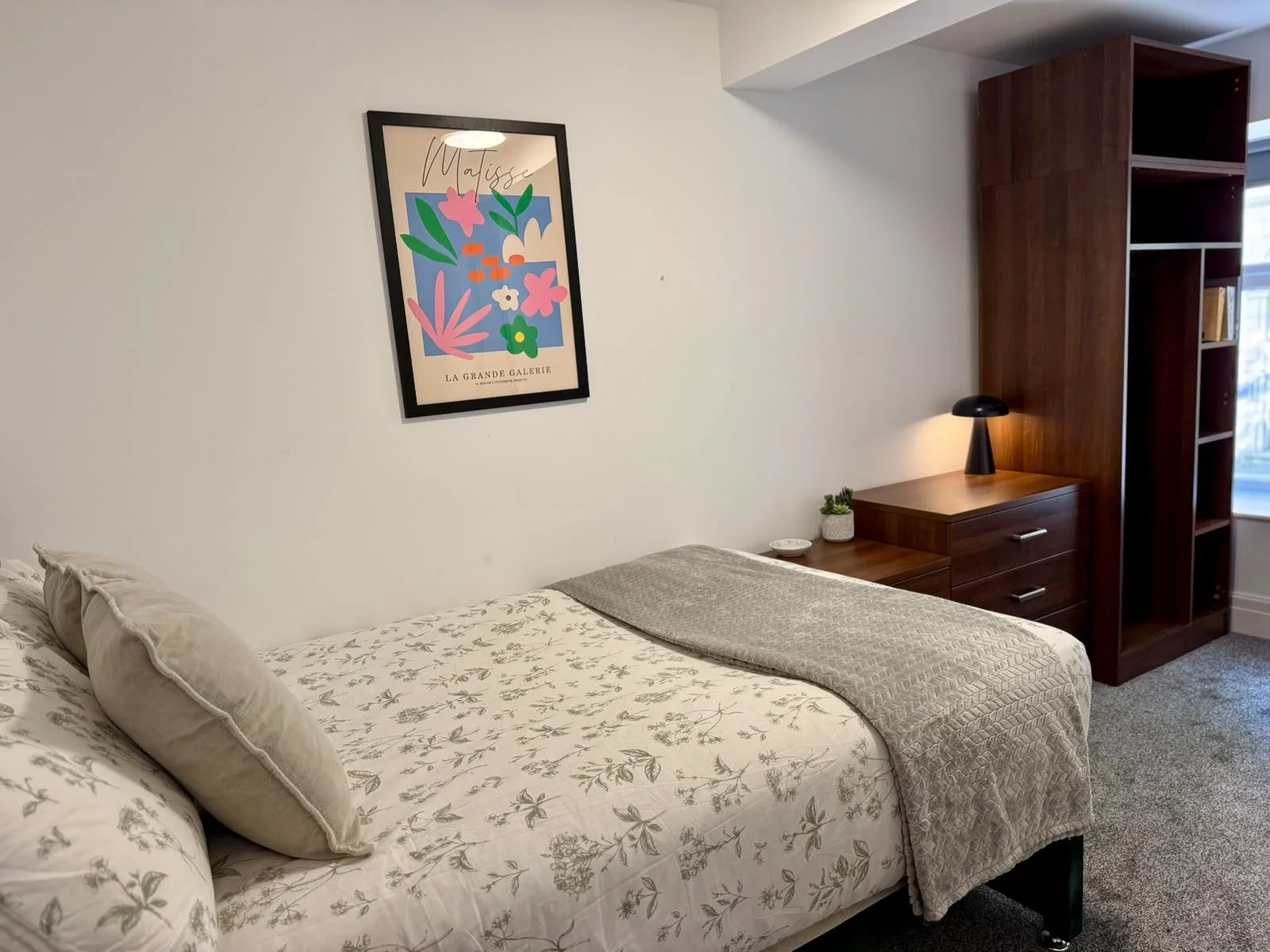A bedroom with a bed featuring floral bedding and beige pillows, a wooden nightstand with a lamp and small plant, a framed abstract floral art piece on the white wall, and a tall wooden bookshelf near a window.