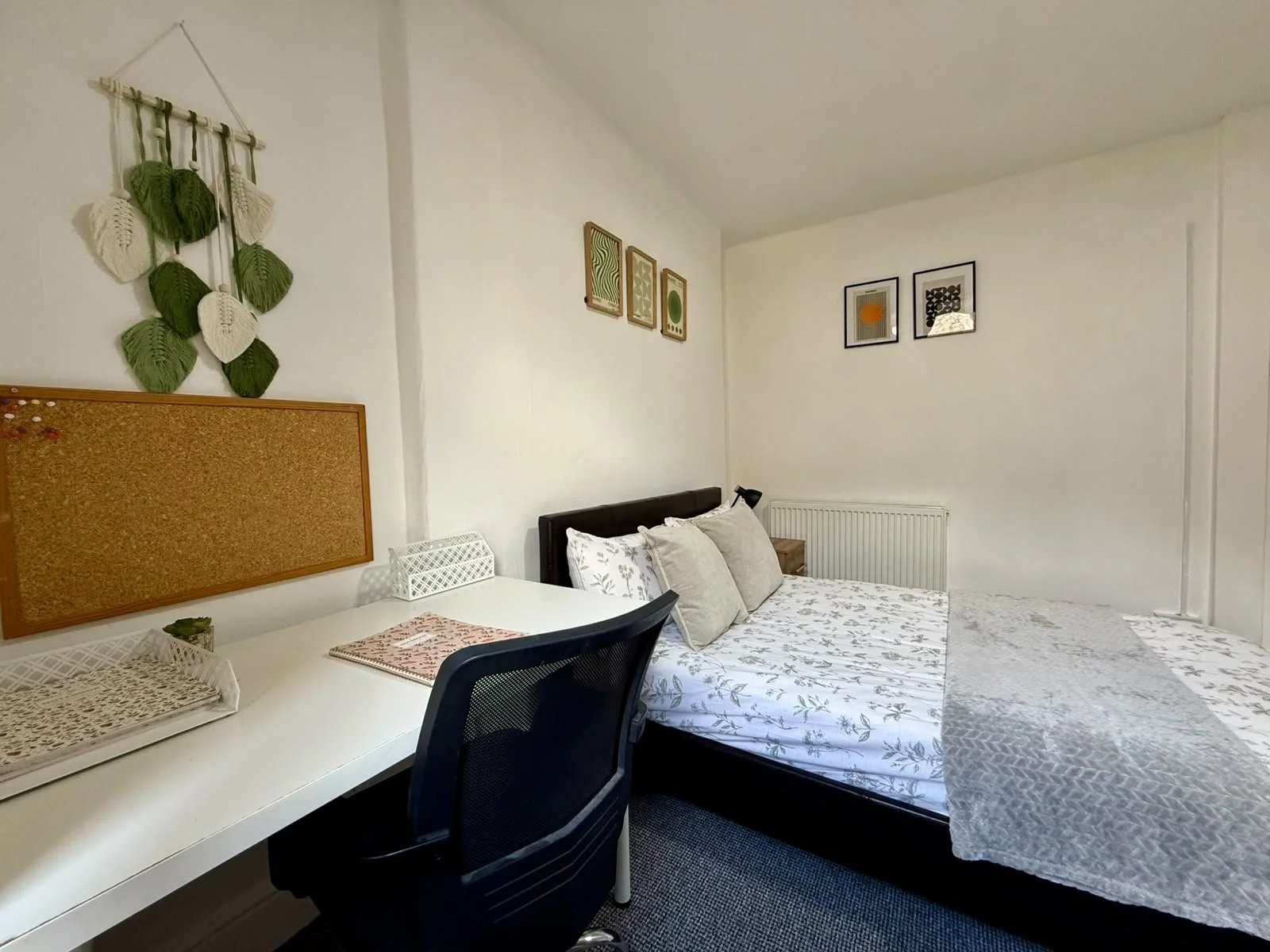 A small bedroom with a bed, desk, and wall art. The bed has beige pillows and a floral bedspread. The desk has a woven tray, a small plant, and a honeycomb patterned box. Wall art includes framed prints and a green and white textile wall hanging.