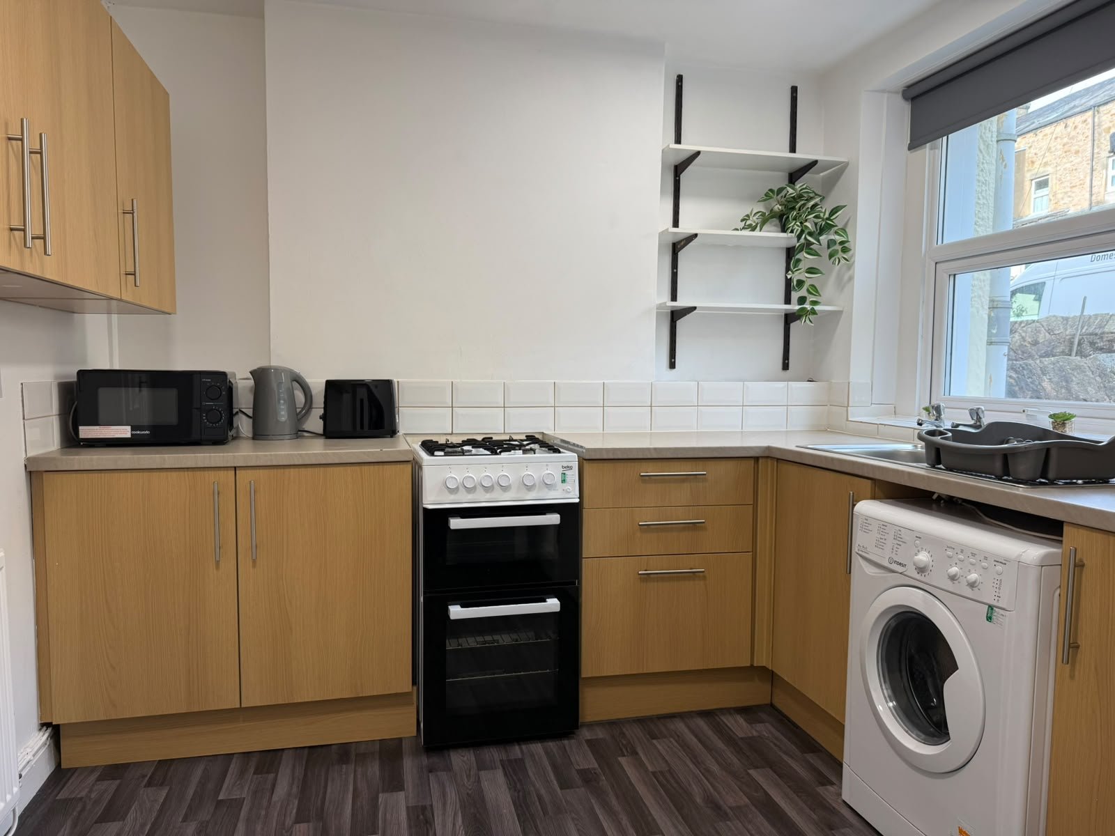 Kitchen with wooden cabinets, white tiled backsplash, black stove, microwave, kettle, toaster, window with grey blind, black sink, and washing machine.