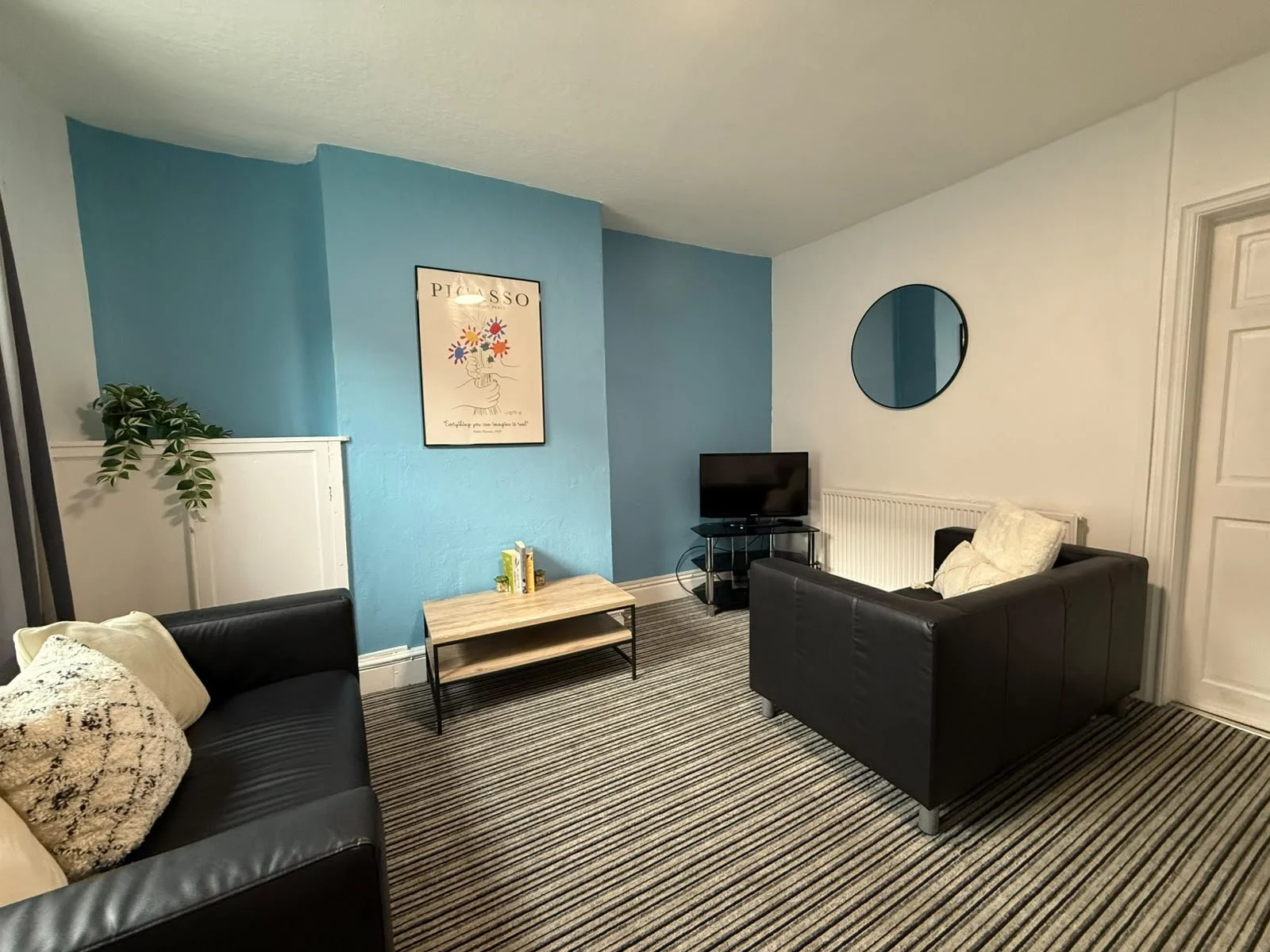 Living room with black sofas, a small TV on a metal stand, a coffee table with books, a round mirror on the white wall, a painting with colorful flowers, a potted plant, and striped carpet.