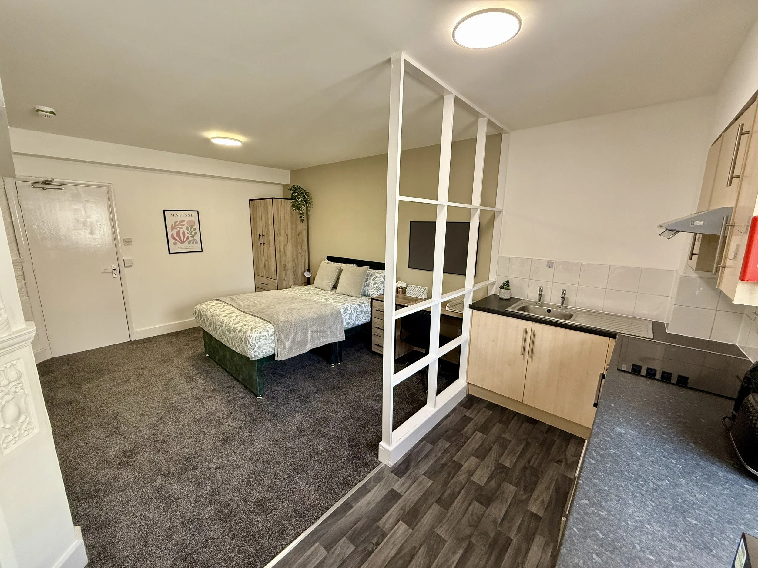 Studio apartment with a bed featuring light-colored bedding, a wooden wardrobe, small bedside table, flat-screen TV on a white grid partition, kitchenette with sink, cabinets, and countertop.