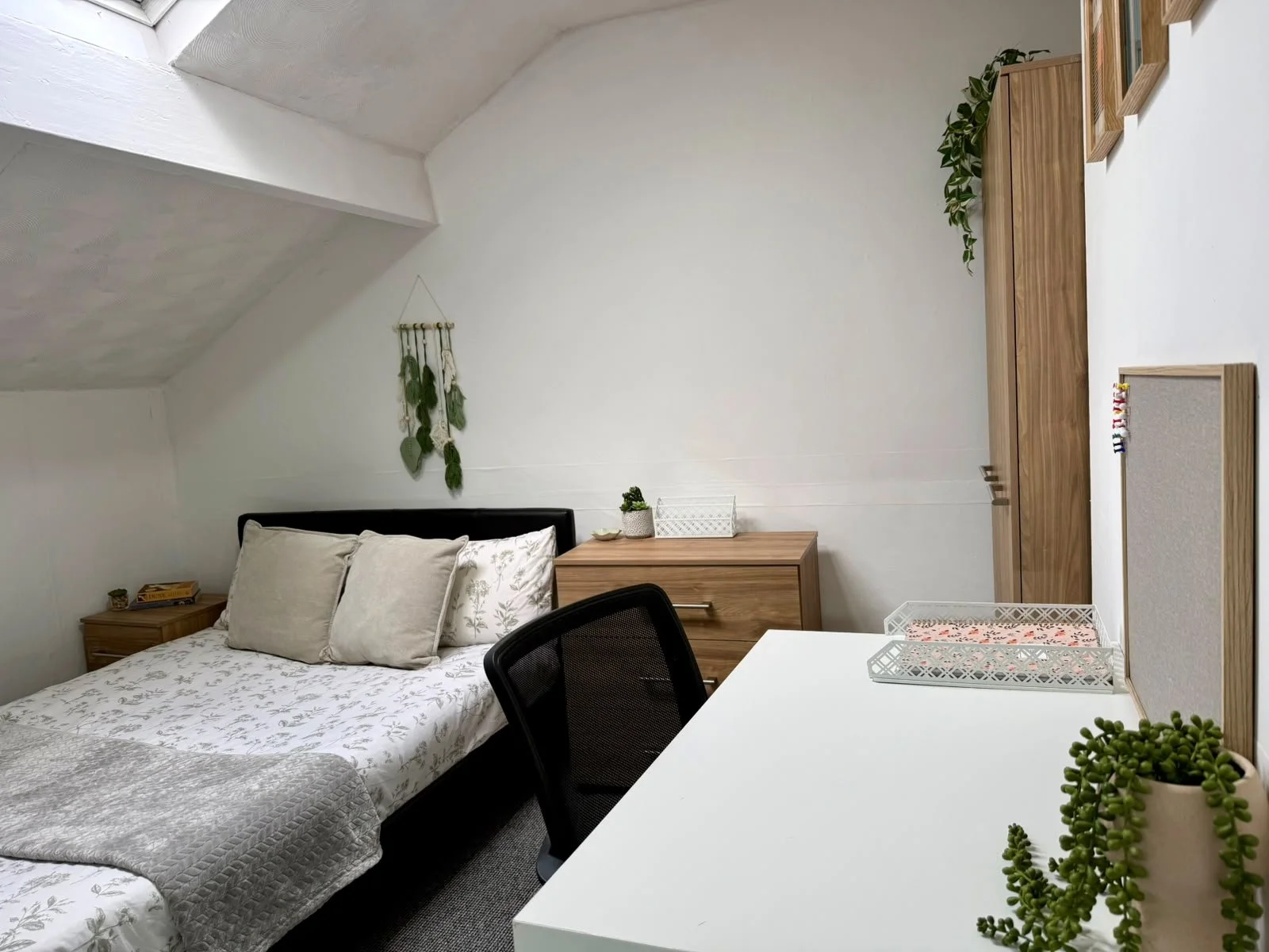 Bedroom with white walls, a bed with beige floral bedding, pillows, a wooden nightstand, and a black chair. There are green plants and hanging decor on the wall, a wooden dresser, and a white desk with a decorative tray and small plant.