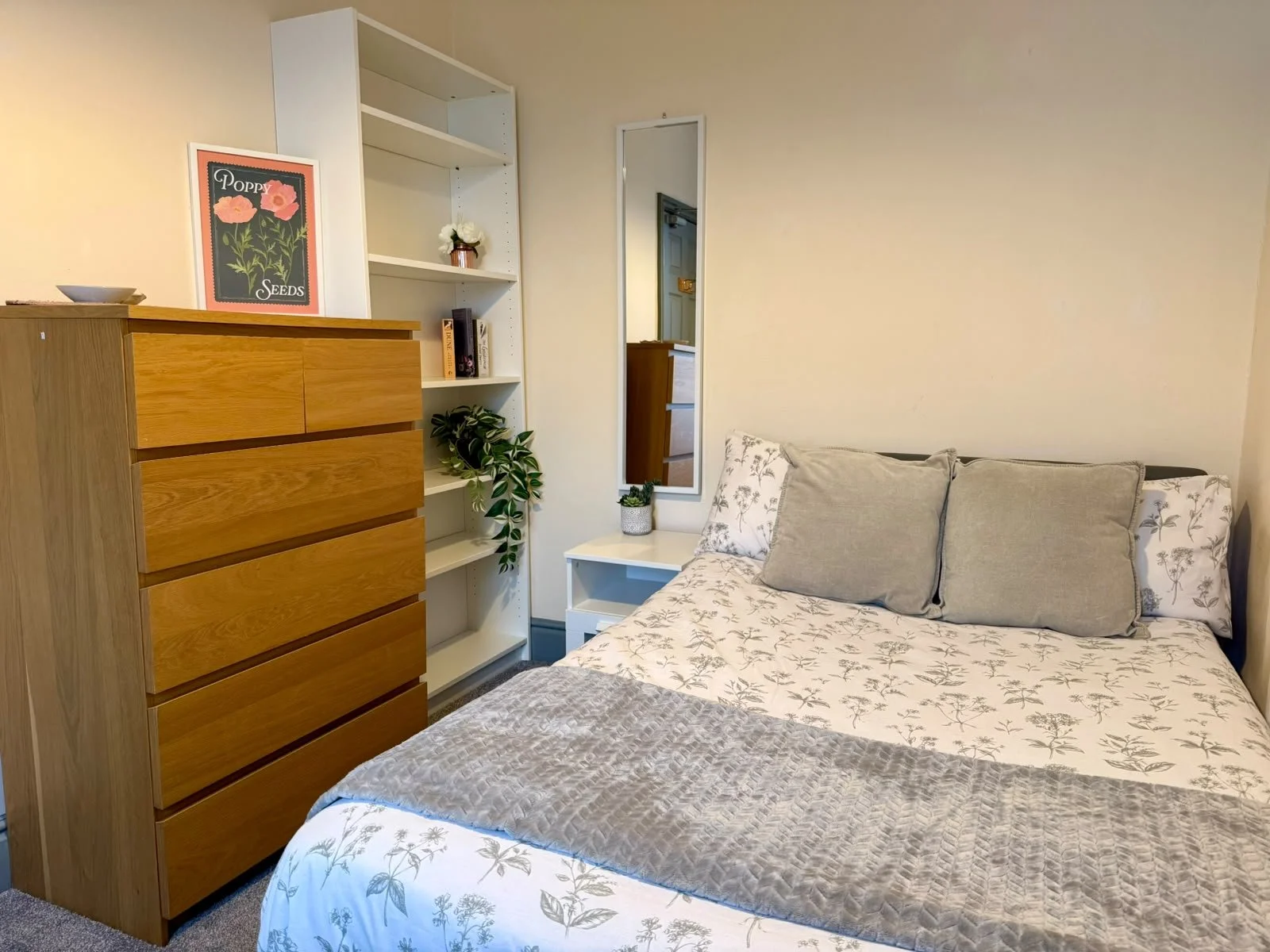 A bedroom with a wooden dresser, a white bookshelf, a bed with floral bedding and pillows, a small white nightstand with a plant, a mirror on the wall, and a beige wall.