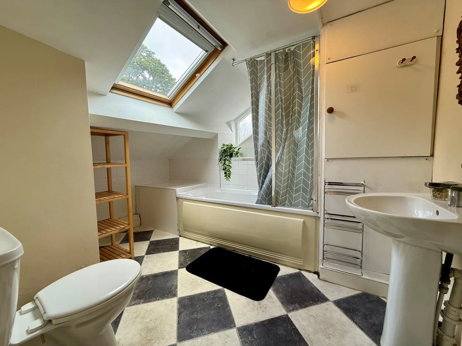 A bathroom with a toilet, a sink, a wooden shelf, a bathtub with a shower curtain, a skylight window, and black and white checkered flooring.
