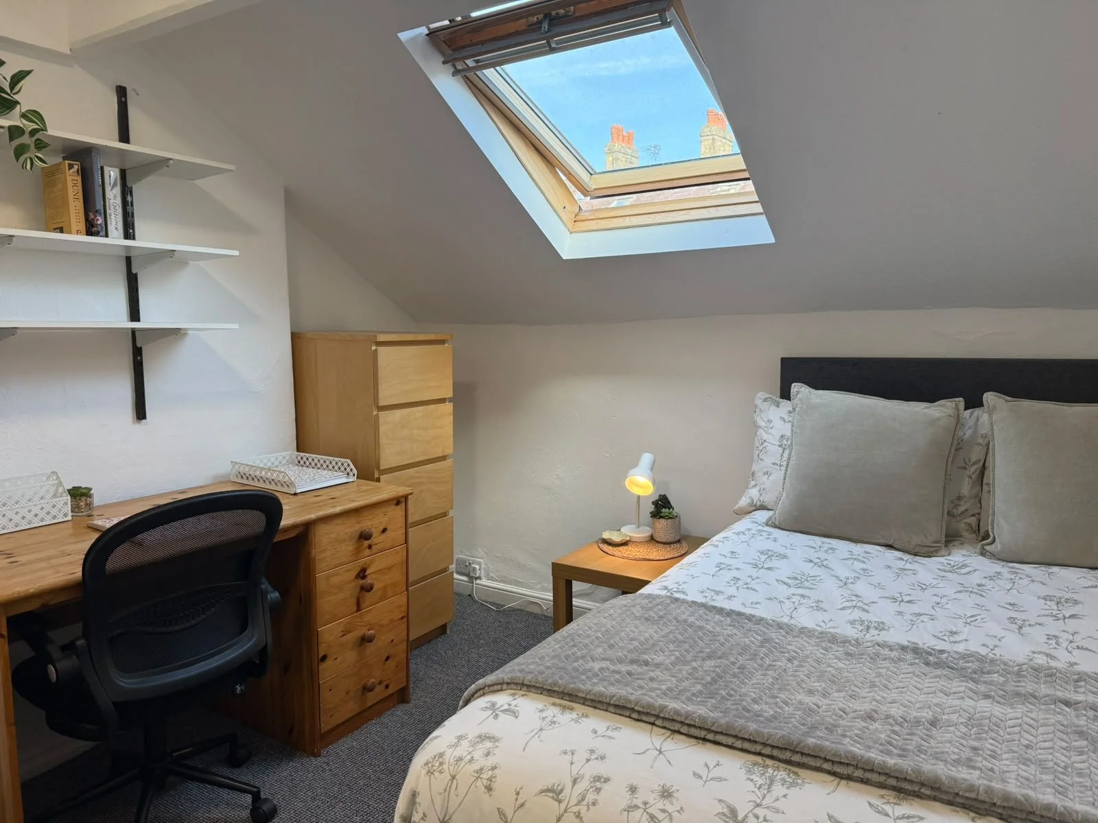 A cozy bedroom with a sloped ceiling and skylight window, a bed with pillows, a bedside table with a lamp, a wooden desk with a chair, a set of wooden drawers, and wall-mounted shelves with books and a plant.