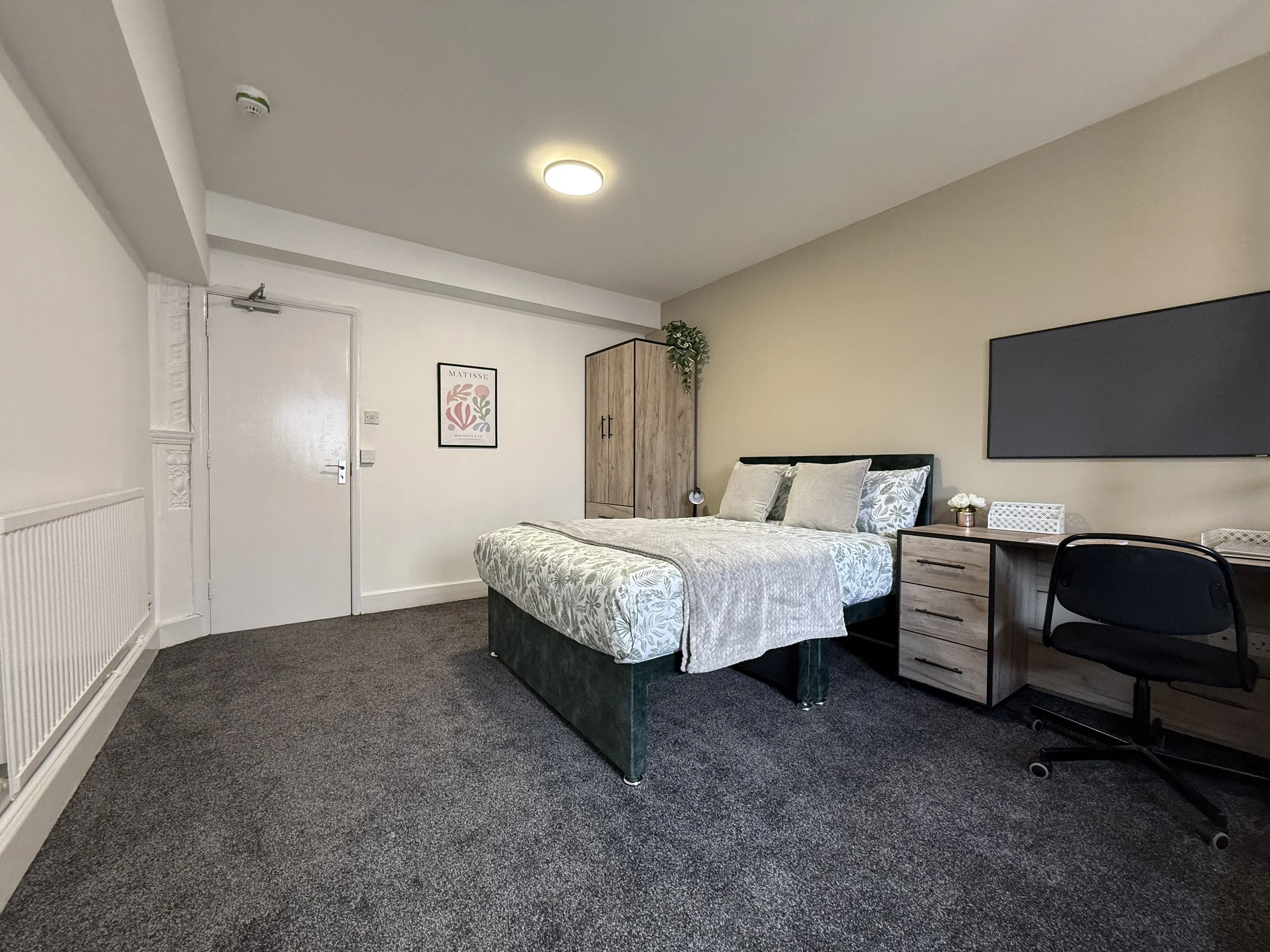 A neatly made bed with white and green floral bedding, two decorative pillows, a wooden nightstand with a small lamp and tissue box, a flat-screen TV on the beige wall, a black office chair, a wooden wardrobe, and a framed art print on the white wall