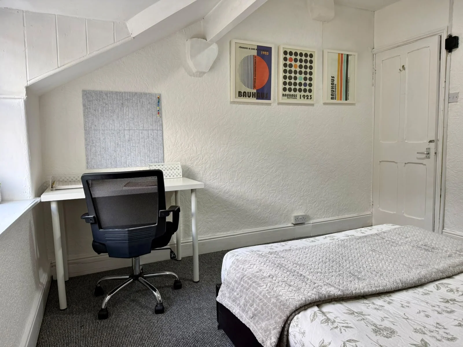 A small bedroom with a white textured wall, black office chair at a white desk, bed with a white and grey patterned blanket, and three framed Bauhaus posters on the wall.