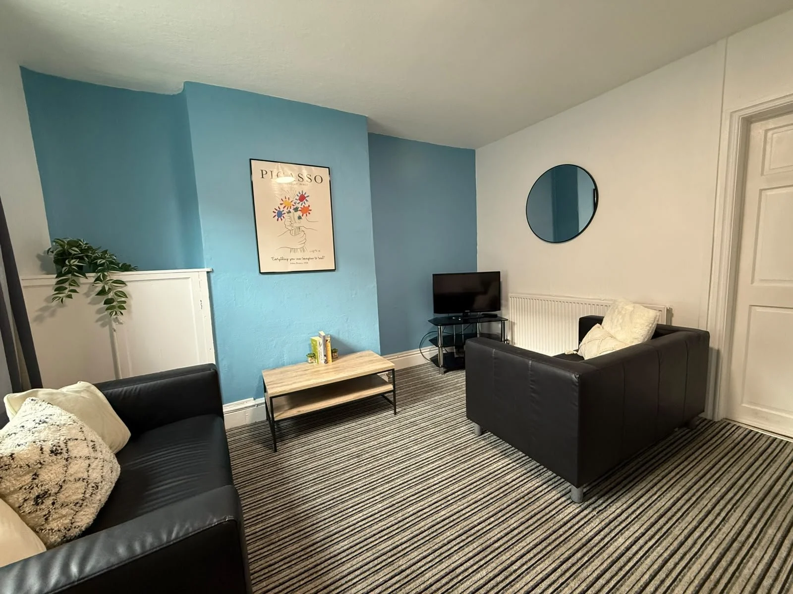 A living room with a black sofa, a light wooden coffee table, a TV on a stand, a black armchair with a white pillow, a round mirror on the wall, a blue accent wall with a poster, and a potted plant.