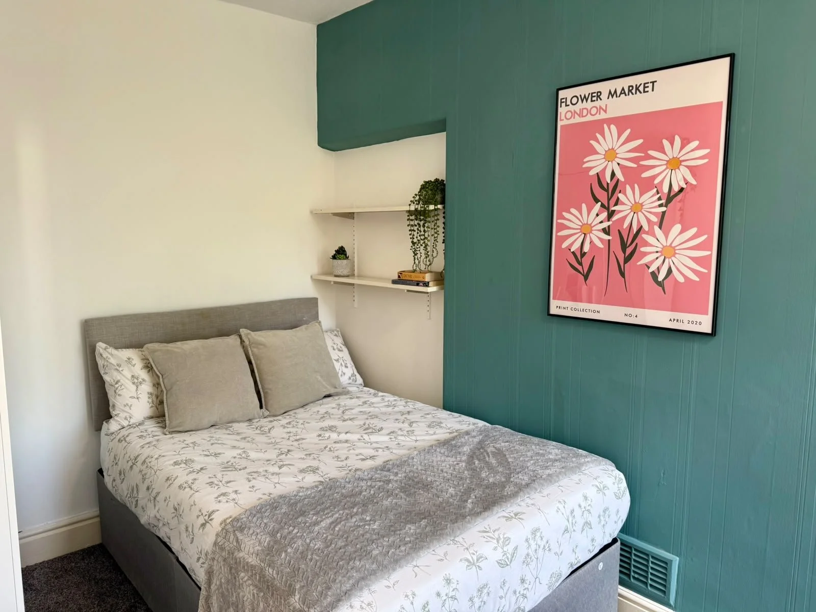 A comfortable bedroom with a neatly made bed featuring gray and white floral bedding and three pillows, viewed against white and teal walls. Two white floating shelves on the teal wall hold small potted plants and books, alongside a large framed art 