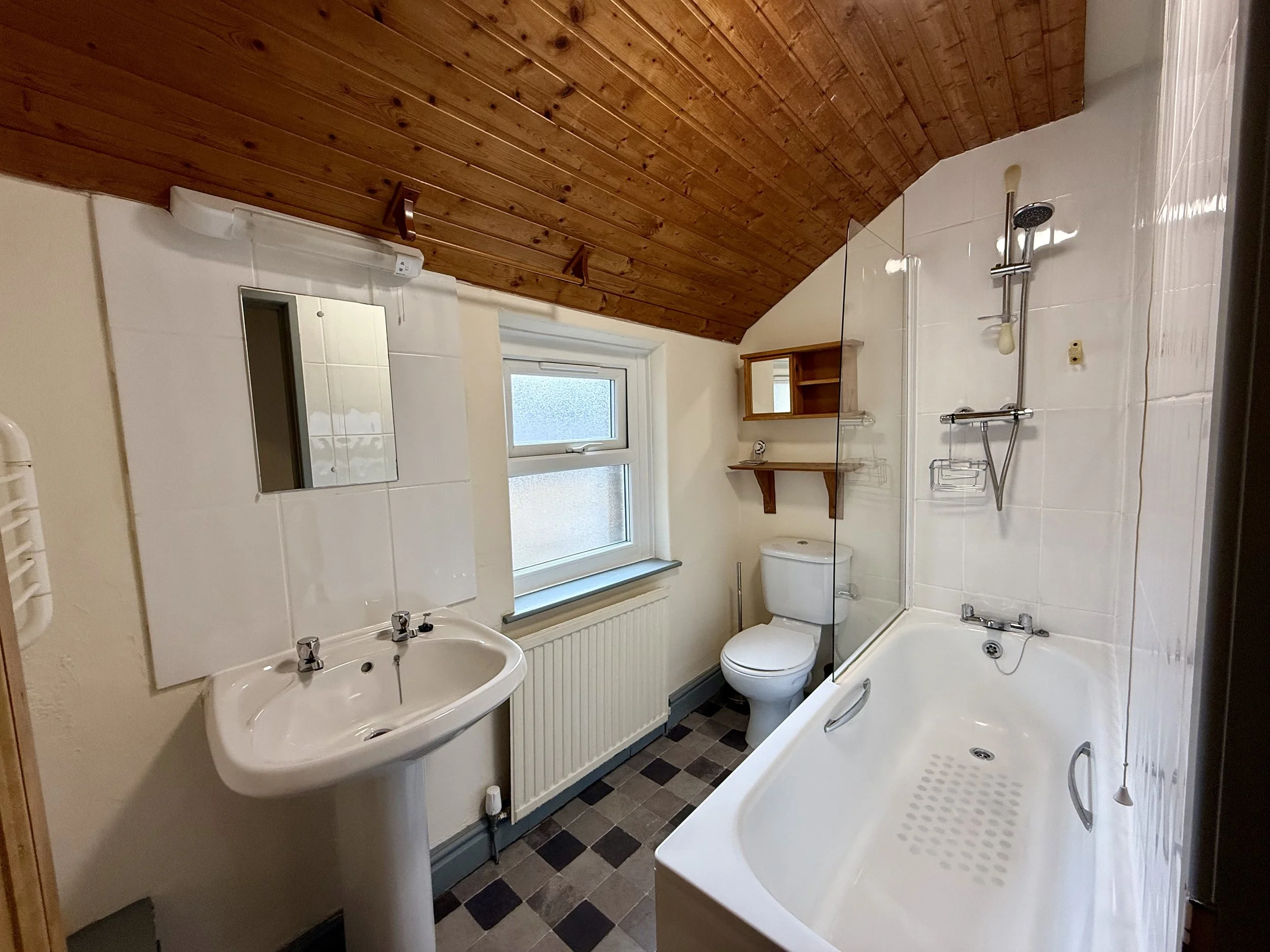Small bathroom with wooden ceiling, white sink, mirror, window, toilet, and bathtub with shower head.