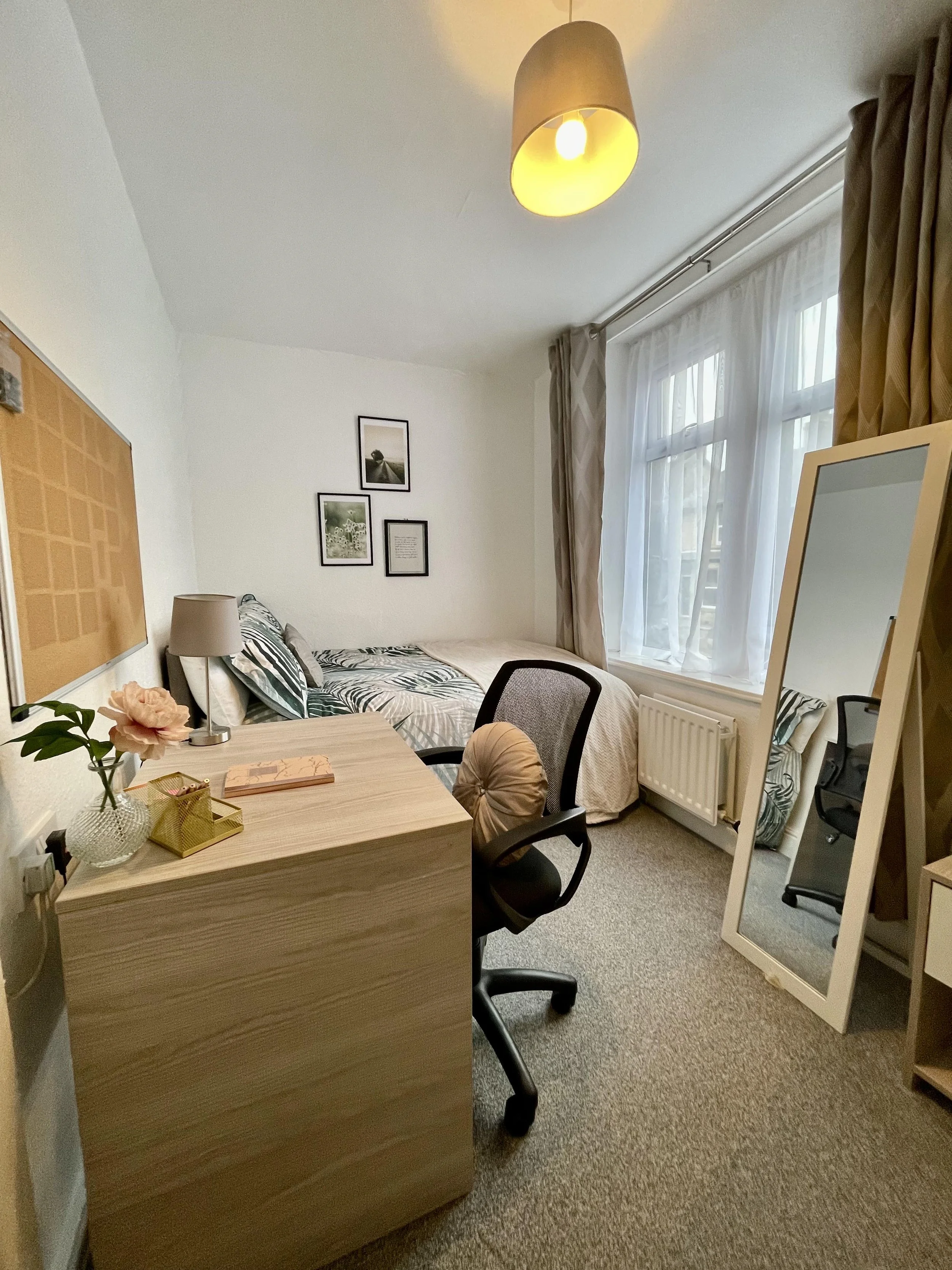 A cozy bedroom with a wooden desk, a black office chair, a large mirror, a bed with patterned bedding, a window with curtains, and wall art.