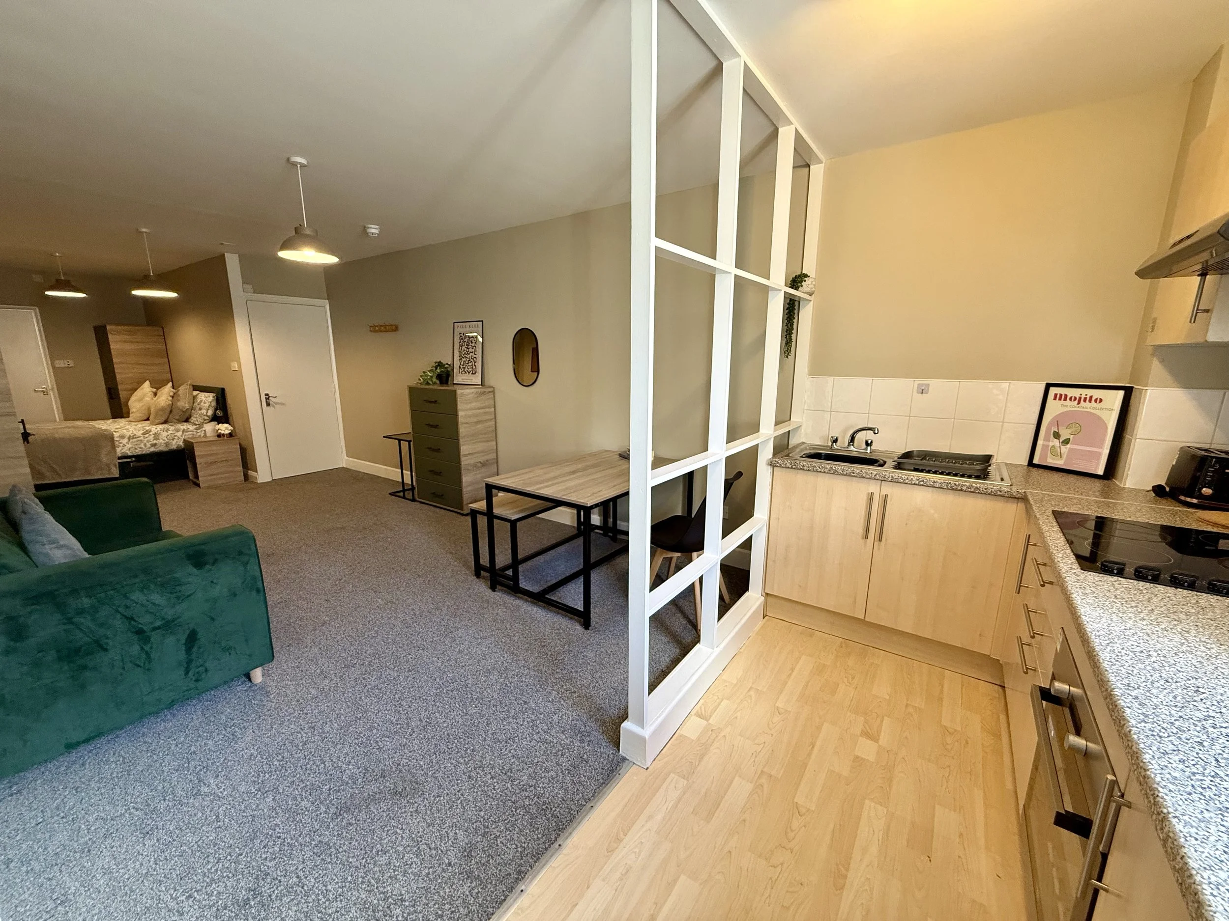 Open-plan living area with kitchen, dining, and lounge space; beige walls, carpeted floor, green sofa, dining table, dresser, bedroom area with bed and pillows in the background, and modern kitchen with light wood cabinets, granite countertops, and s