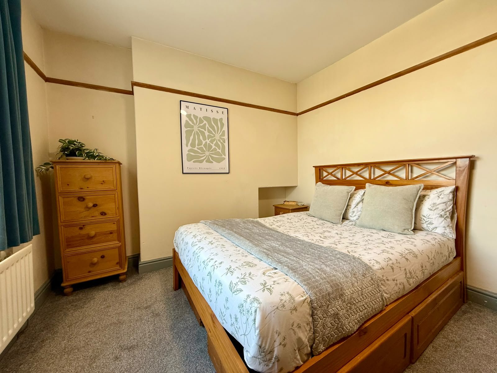 A cozy bedroom with a wooden bed frame, floral bedding, and matching pillows, a wooden dresser with a green plant on top, a bedside table with a clock, and a framed botanical print on a light-colored wall.