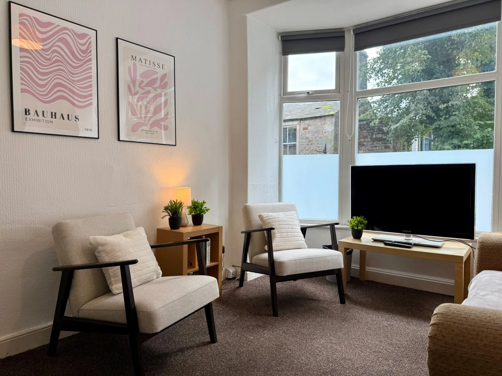A cozy living room with beige armchairs, a wooden side table with plants and a lamp, a black TV on a wooden stand, large windows with views of trees and an old brick building, and framed artwork on the wall.