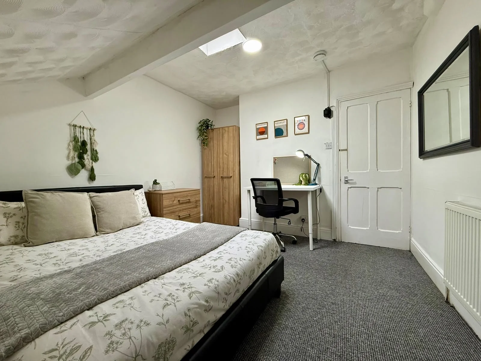 A cozy bedroom with a bed, a wooden wardrobe, a small desk with a black office chair, decorative wall art, and a hanging wall decoration. The room has white walls, a gray carpet, and a sloped ceiling with a skylight.